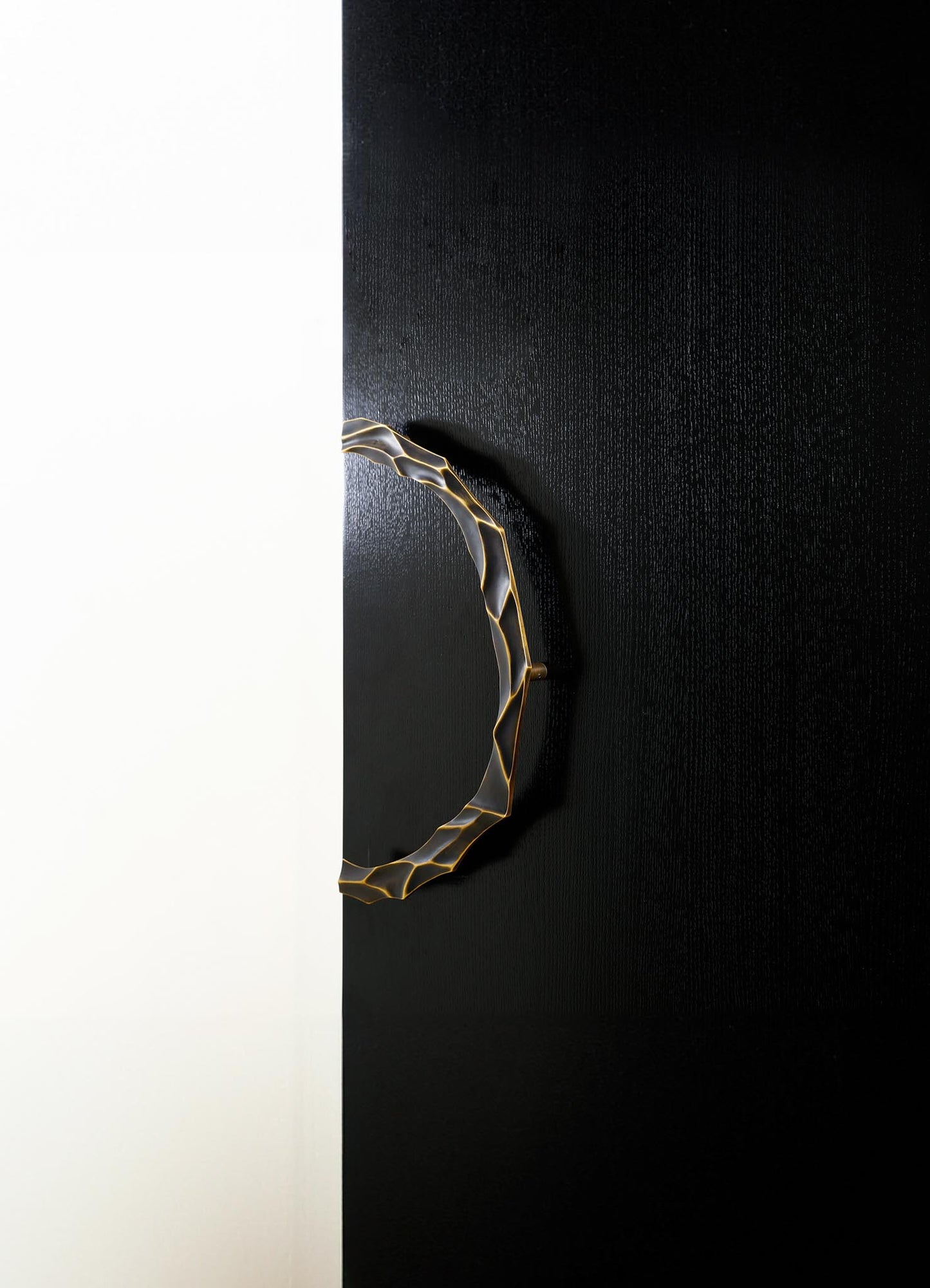 A contemporary decorative mirror with a sculptural brass frame featuring an organic, irregular pattern, mounted on a black textured wall. The mirror has a round shape and a metallic gold finish.