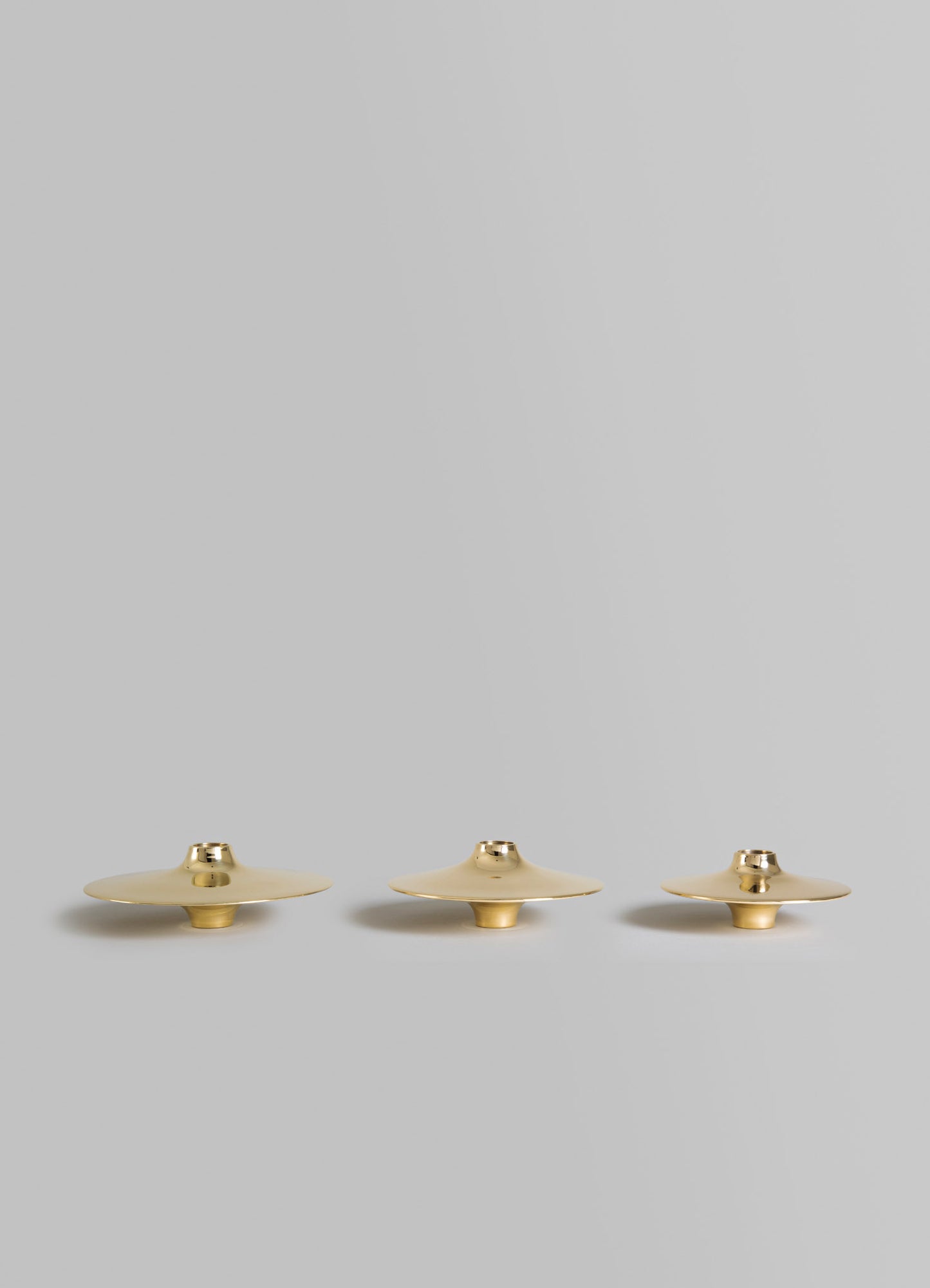 Set of three brass candles holders with a flat, disc-like shape and a smooth, polished finish, arranged in decreasing size on a light gray background.