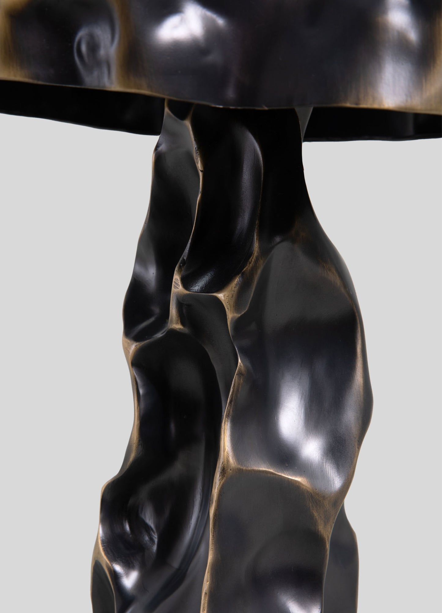 A close-up of a sculptural table base made of dark, polished metal with organic, flowing curves and a textured, irregular surface, supporting a smooth, round tabletop.