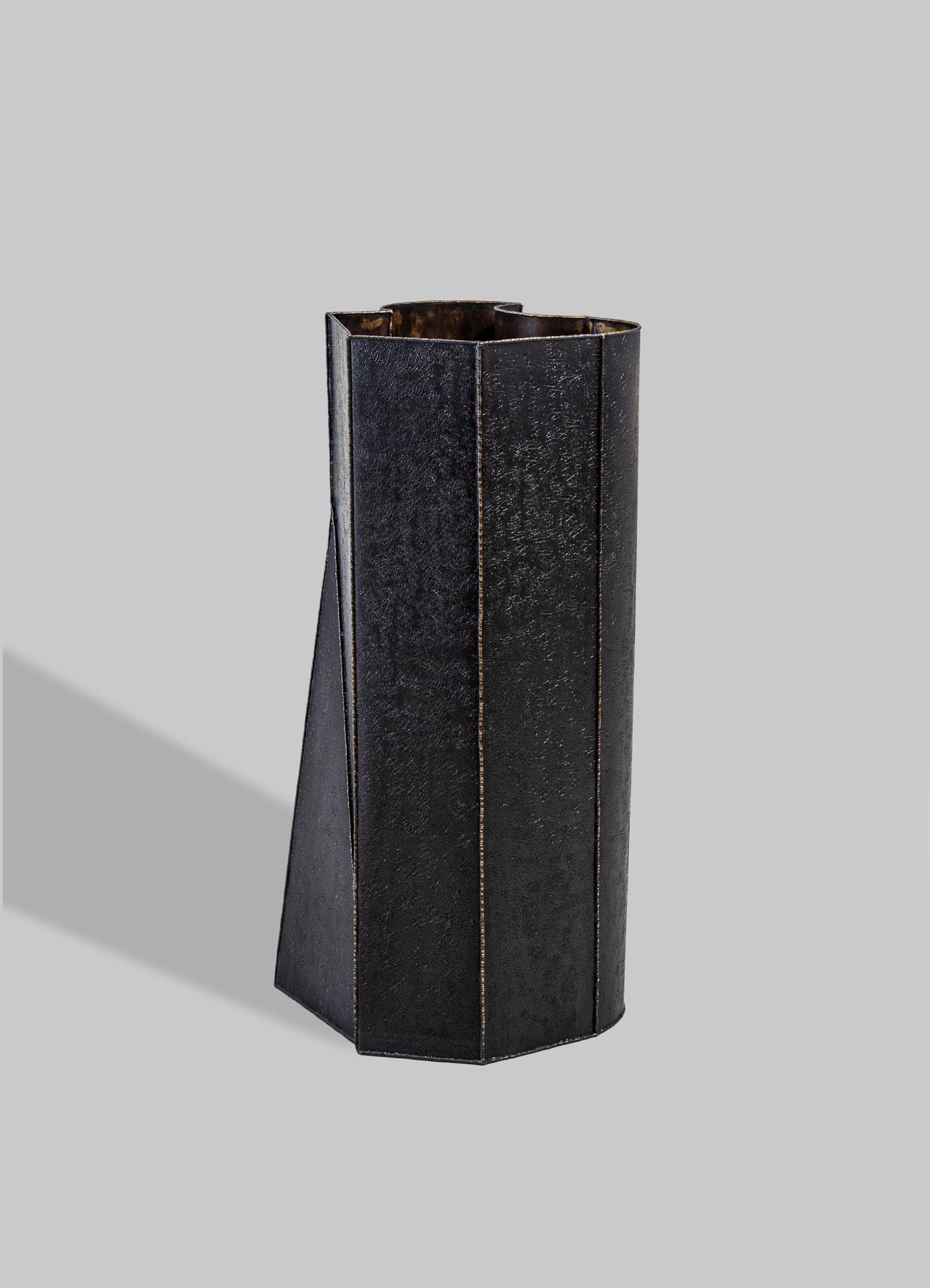 A tall, black metal vase with a textured surface and geometric, angular lines, set against a light gray background.