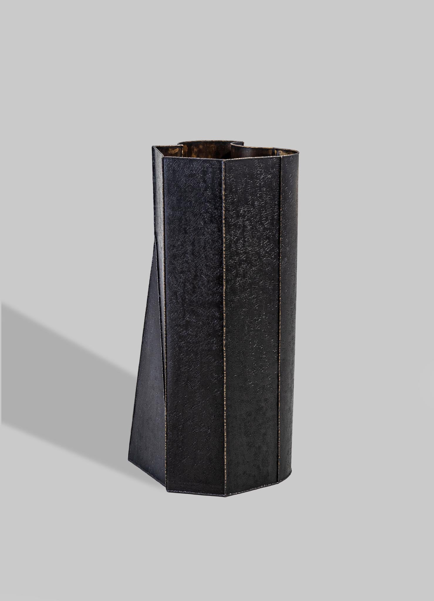 A tall, black metal vase with a textured surface and geometric, angular lines, set against a light gray background.