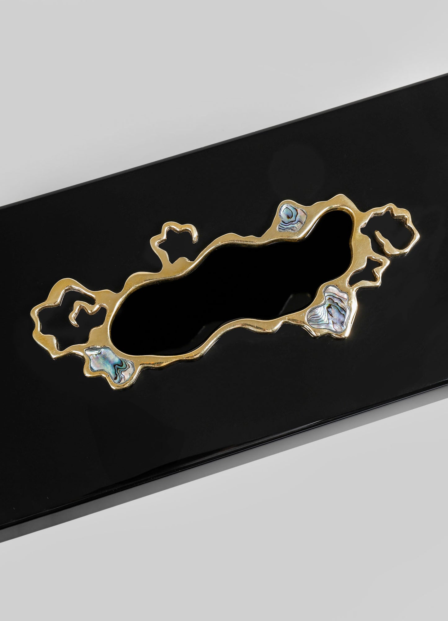 A decorative brass tray with an irregular, organic shape accented by four abalone shell inlays, placed on a black surface. The tray has a modern, handcrafted style with a polished gold finish.
