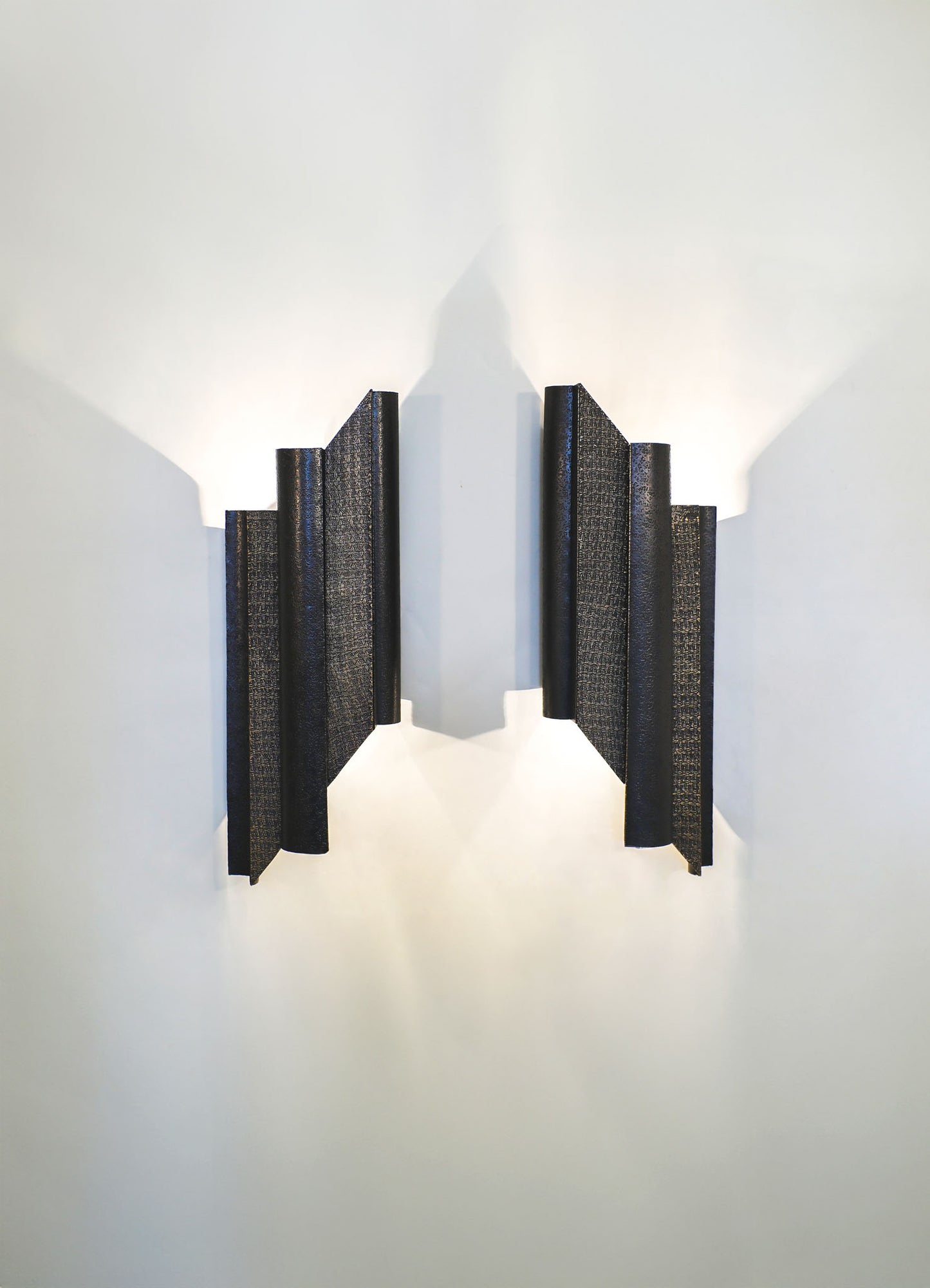 Wall-mounted light fixture with rectangular, textured black panels and a geometric design, casting light and shadows on a white wall. The fixture has a contemporary style with a focus on clean lines and modern aesthetics.
