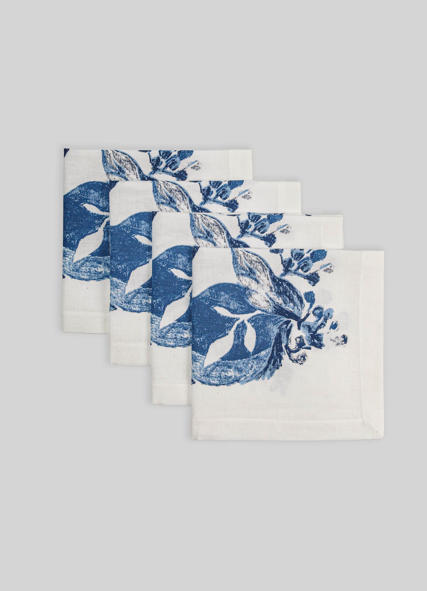 Linen napkins with a blue botanical print featuring leaves and small flowers on a white background, arranged in a fan shape against a light grey backdrop.