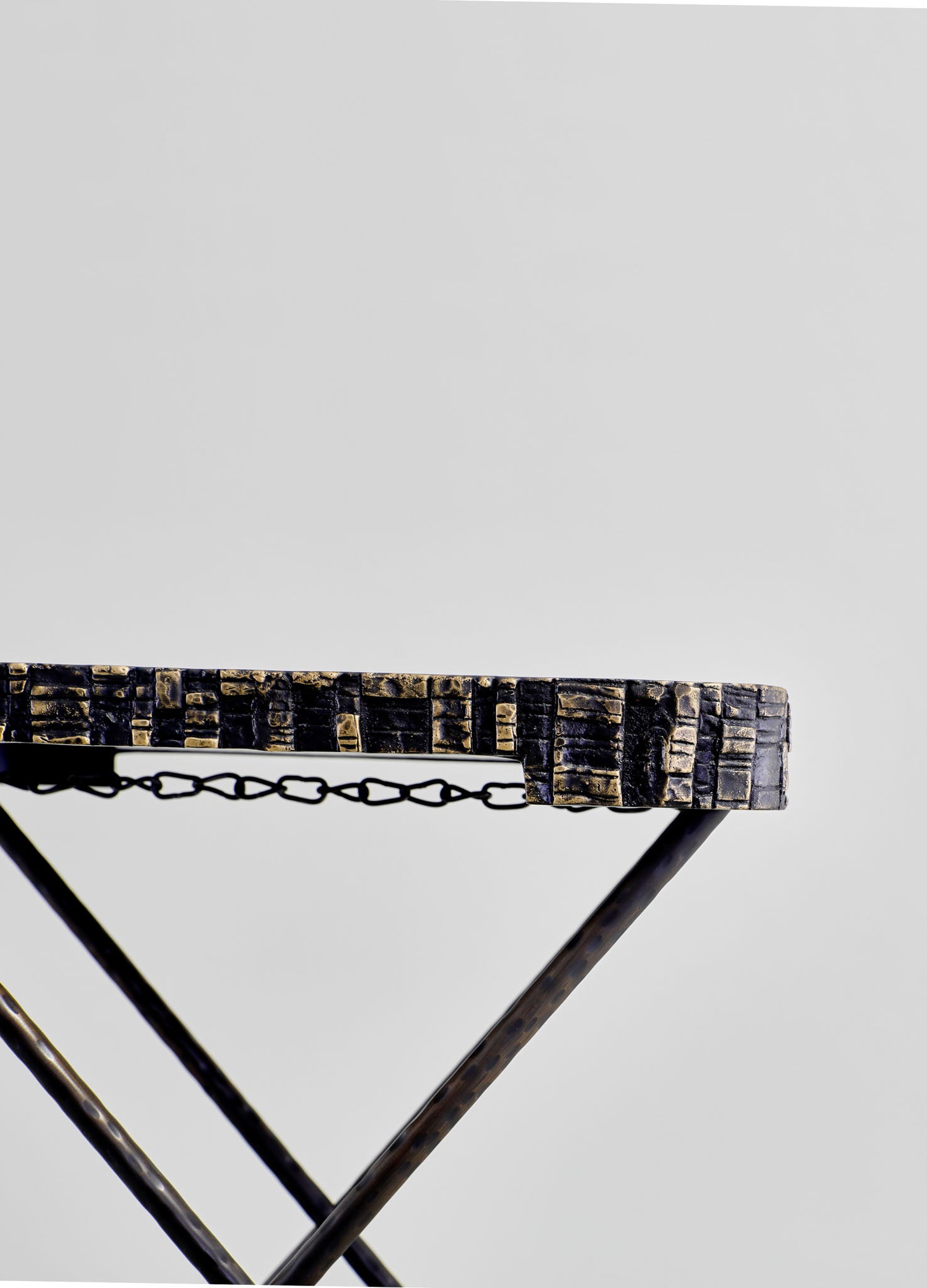 A metal table with a textured, dark finish and a chain detail along the edge, supported by angled legs in a minimalist style. The table has a contemporary design with industrial accents, set against a plain light background.