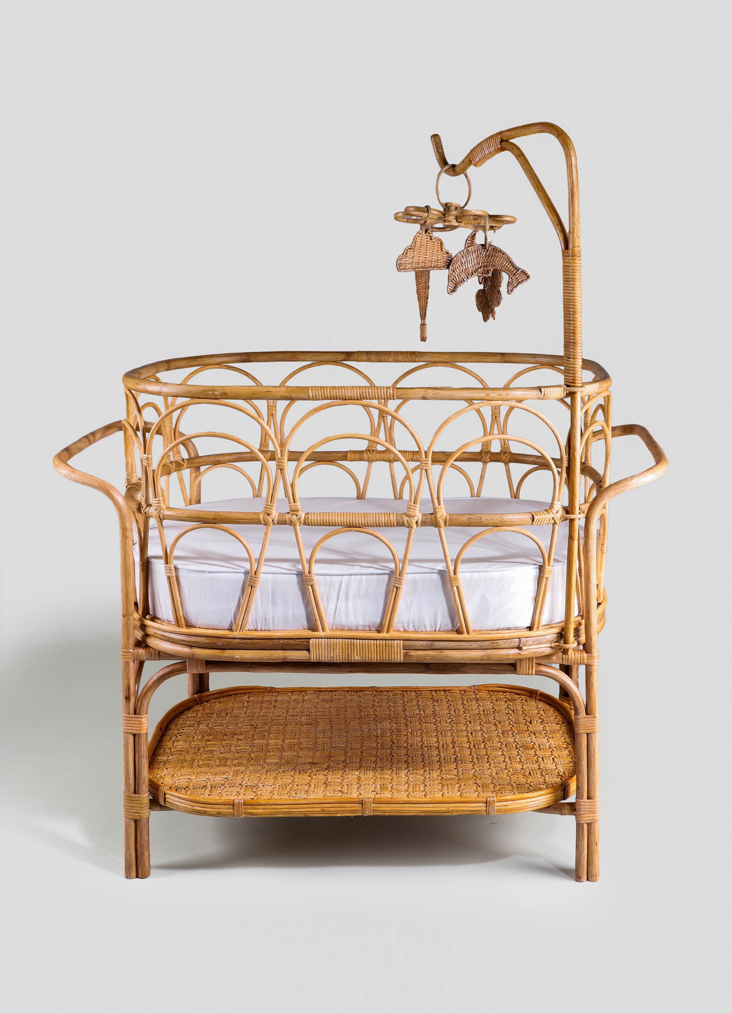 Rattan bassinet with a woven bottom shelf and a decorative upper frame, featuring a scalloped design and an attached mobile with wicker animal motifs. The natural wood finish emphasizes a handcrafted, rustic style suitable for a nursery or child's space.
