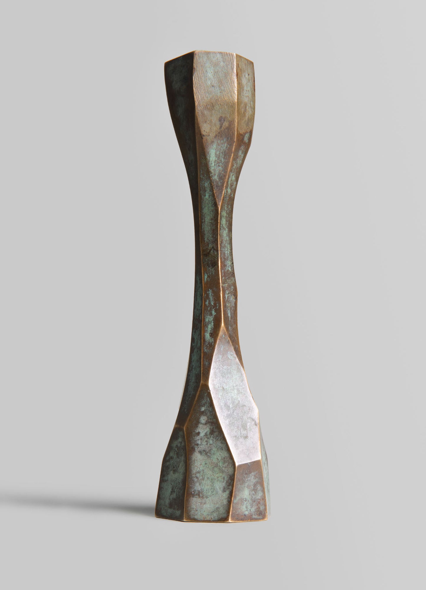 A sculptural vase made of metal with a textured, oxidized finish in shades of bronze and green. The vase features a tall, slender, twisted form with faceted geometric surfaces, embodying a contemporary and artistic style.