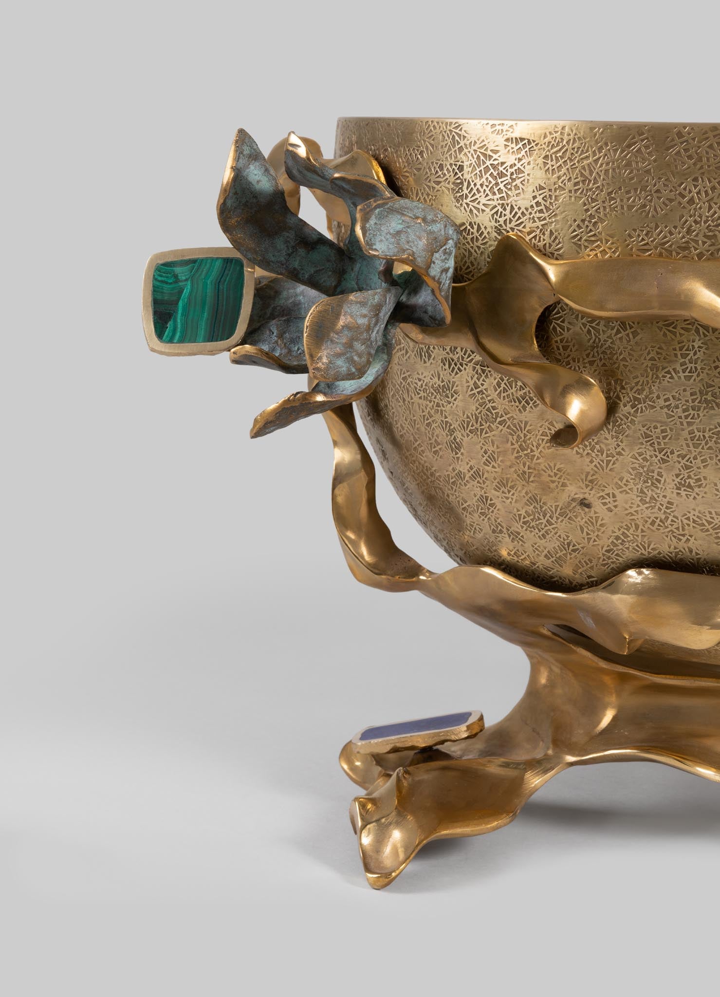 Decorative metal bowl with textured gold finish and organic, flowing edges, featuring a sculptural flower motif with metallic leaves and a gemstone accent in green.