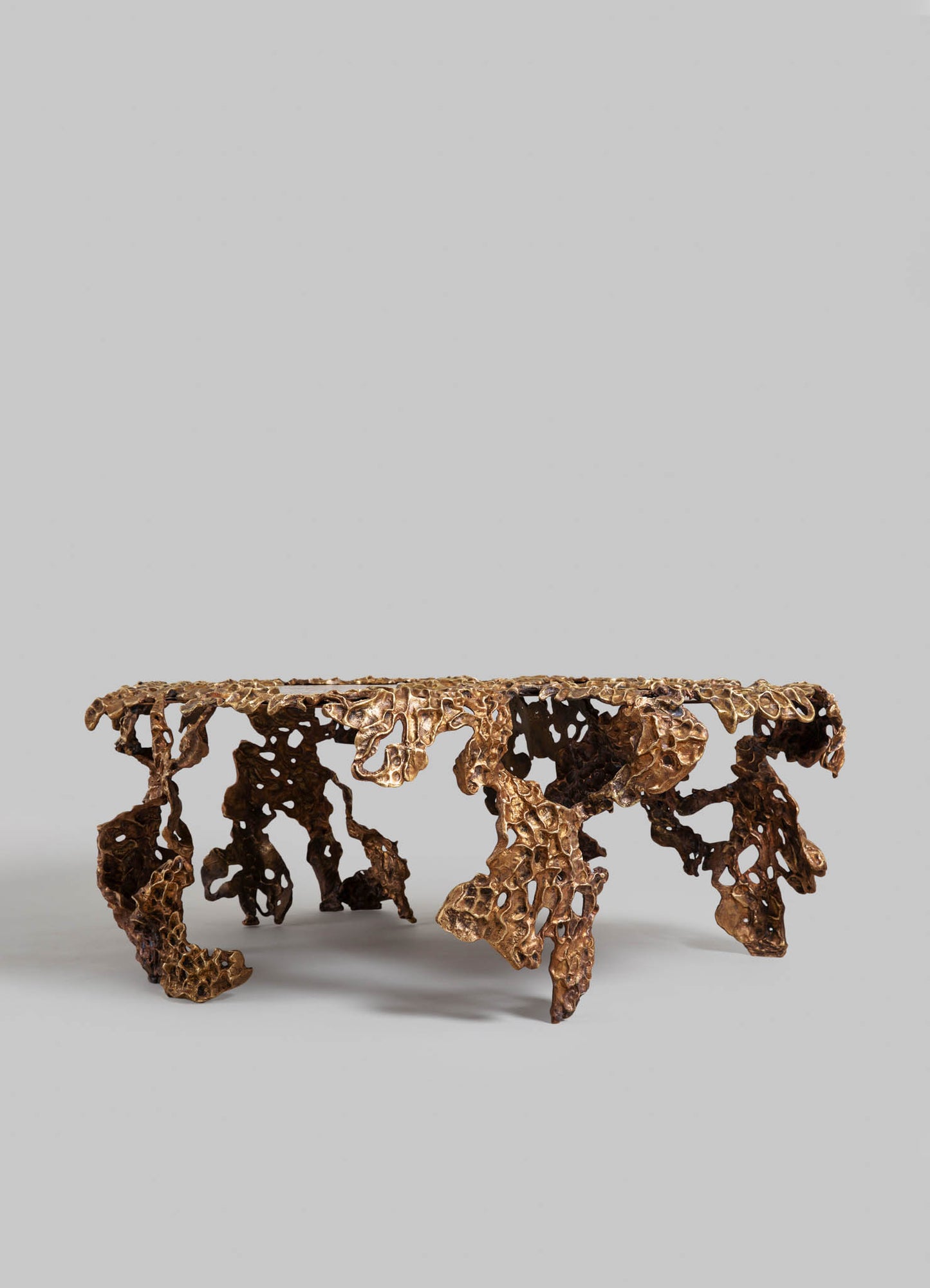 A bronze table with a textured, organic design featuring irregular, porous surfaces and a natural stone-like appearance. The structure exhibits a handcrafted style with a metallic finish, highlighting intricate details and a contemporary aesthetic.