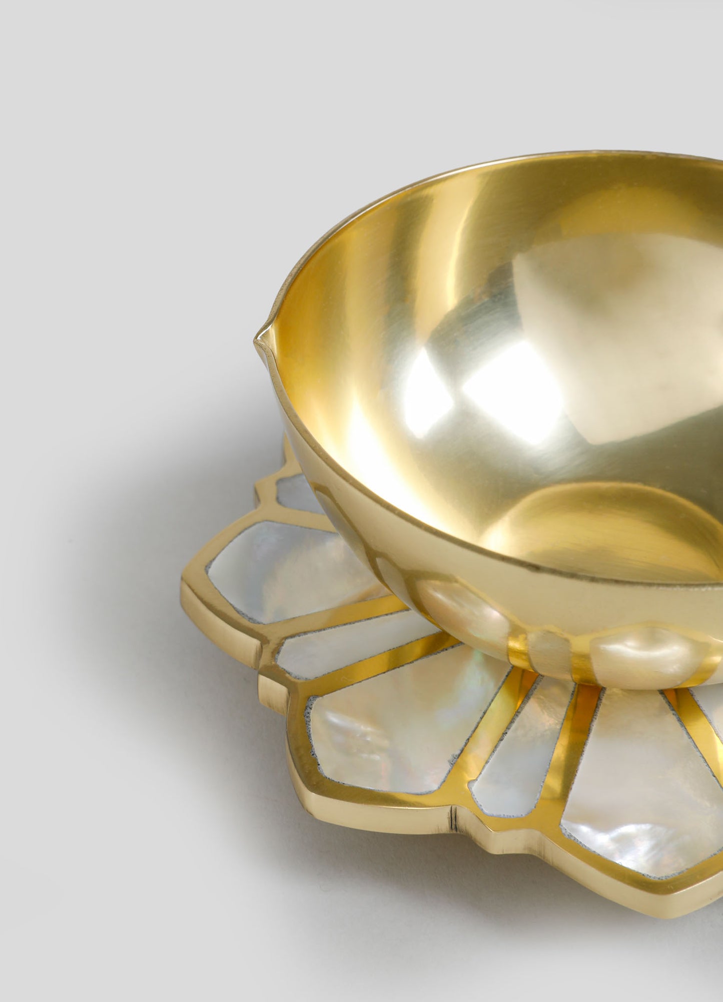 A brass bowl with a smooth, polished interior and a scalloped edge sits atop a decorative base featuring geometric cutouts and mother-of-pearl inlay, creating a contrasting iridescent effect.