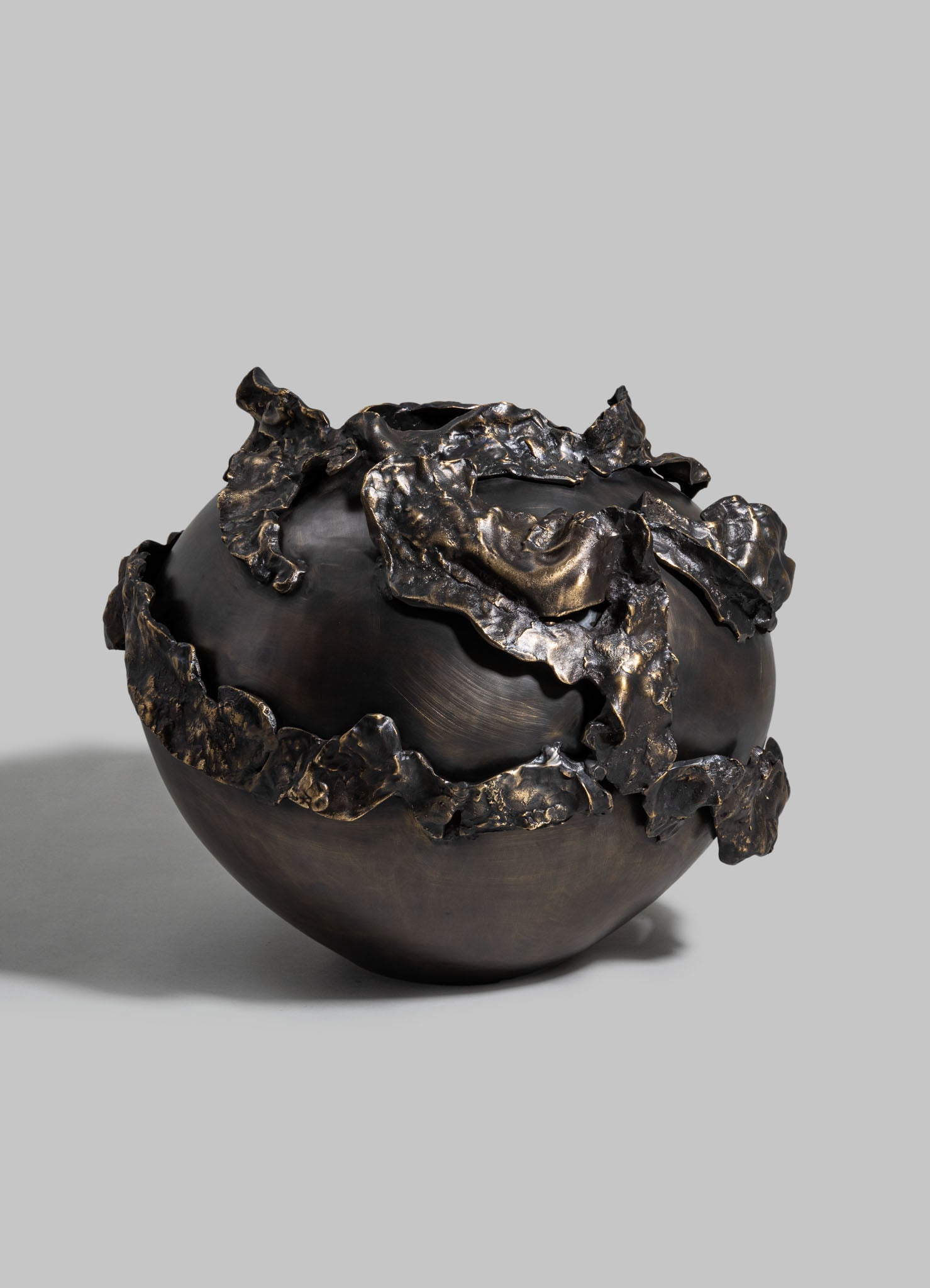 A black ceramic bowl with an organic, rounded shape, featuring textured metallic bronze accents that resemble crumpled or melted metal draped over the surface. The design combines contemporary and sculptural elements with a metallic finish.
