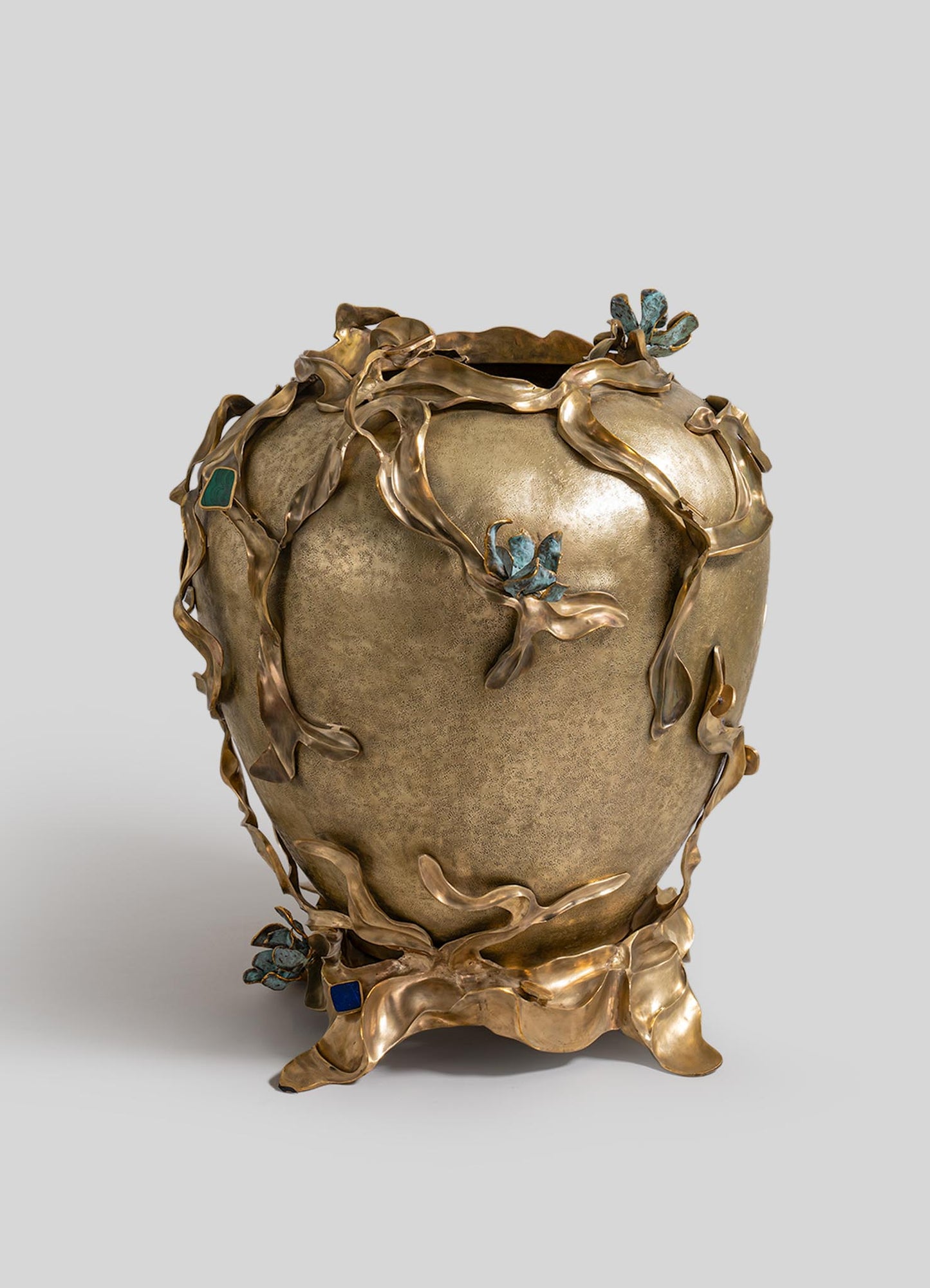 A decorative ceramic vase with a textured beige surface, accented by sculptural metal floral and leaf motifs in bronze with blue patina details, creating an organic, handcrafted appearance.