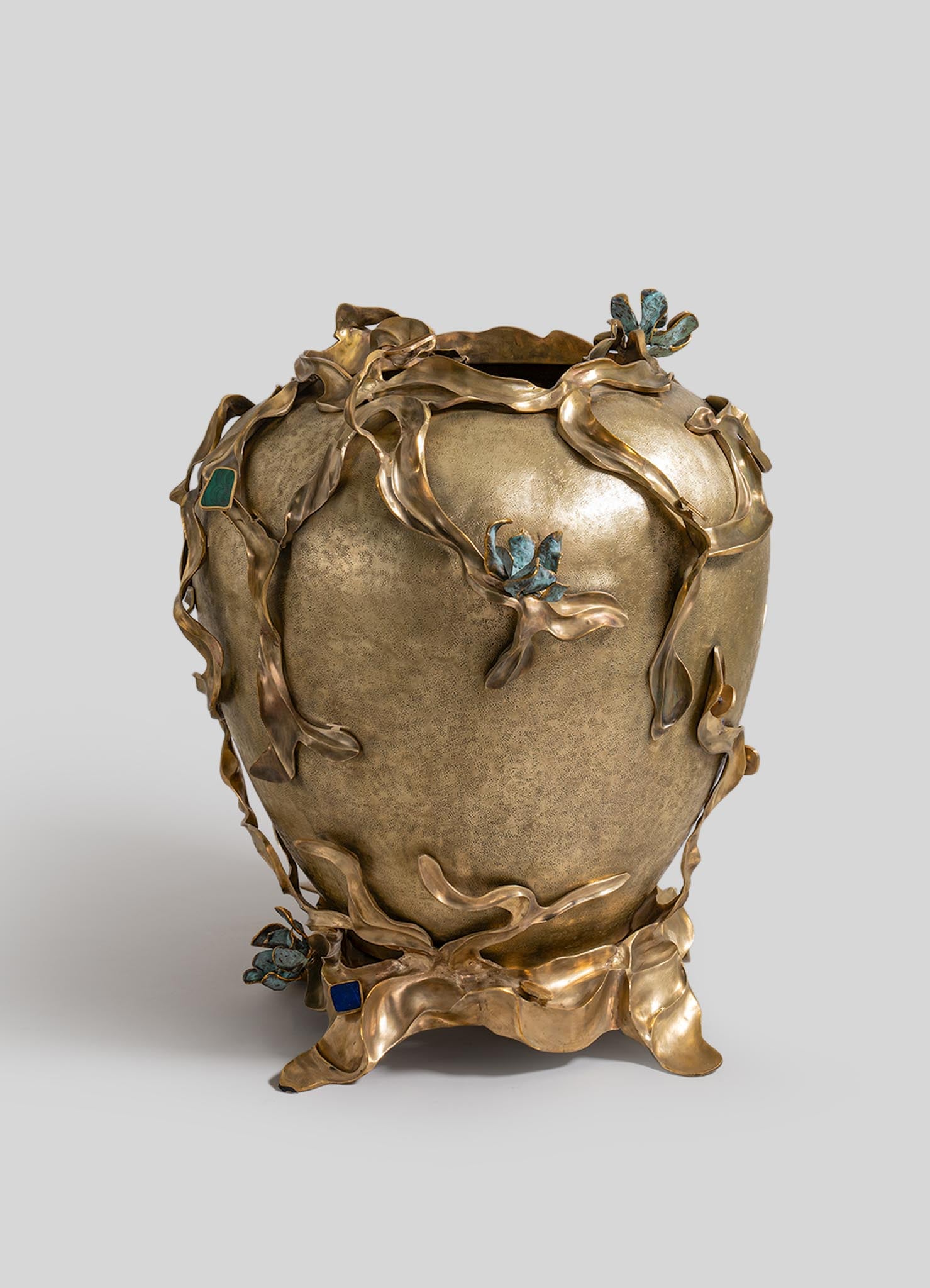 A decorative ceramic vase with a textured beige surface, accented by sculptural metal floral and leaf motifs in bronze with blue patina details, creating an organic, handcrafted appearance.
