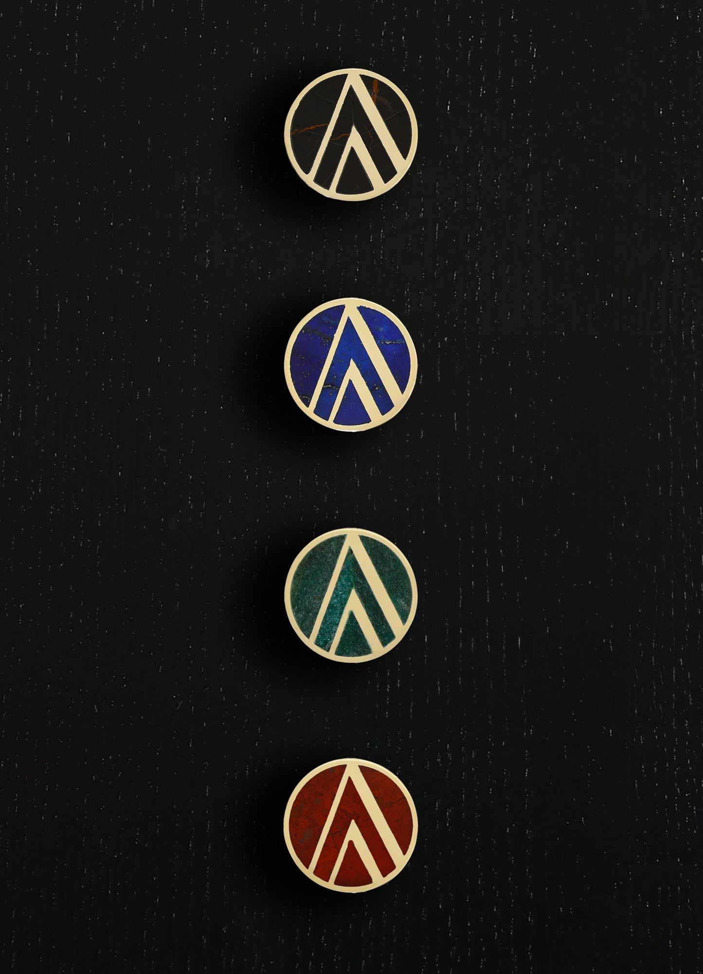 Set of four round enamel pins with geometric designs in various colors including beige, blue, green, and red, featuring triangular and linear motifs on a black background.