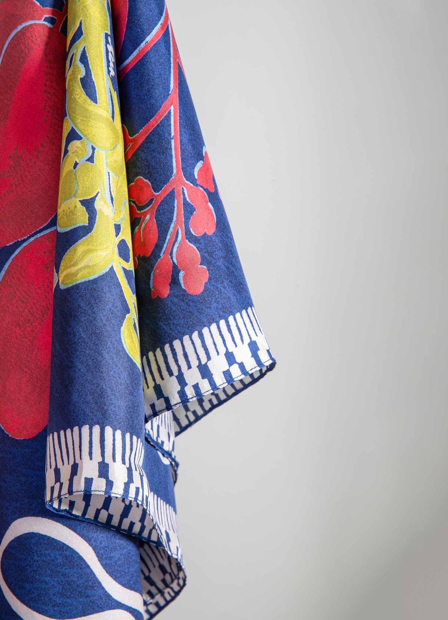 Fabric with a floral pattern in bold red, yellow, and blue colors, featuring botanical motifs and geometric border details. The material appears to be lightweight, possibly cotton or linen.