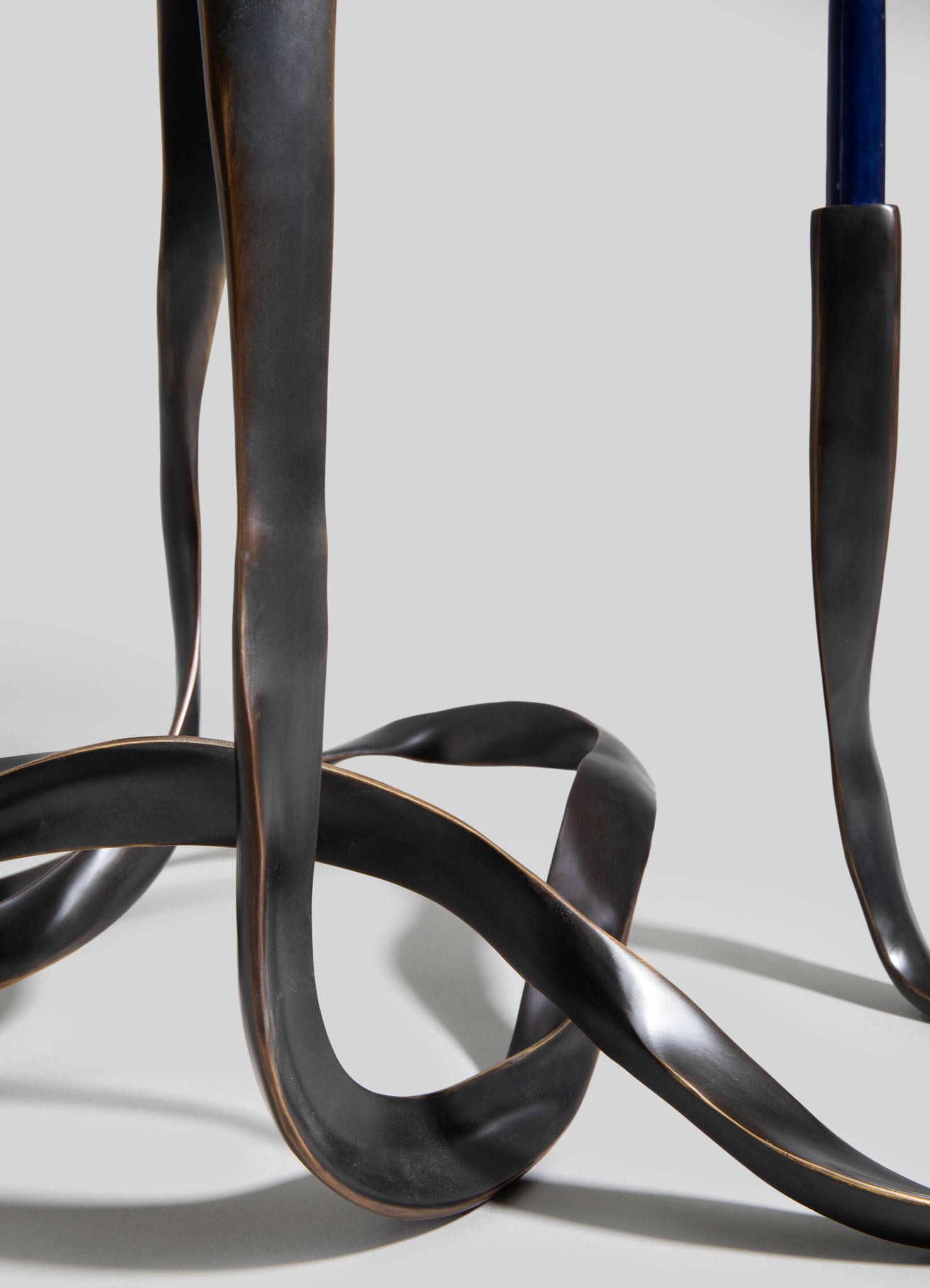 Close-up of a sculptural metal stand with twisted, organic-shaped legs in dark bronze finish, featuring hints of metallic sheen. The design emphasizes abstract, flowing lines with a contemporary style.