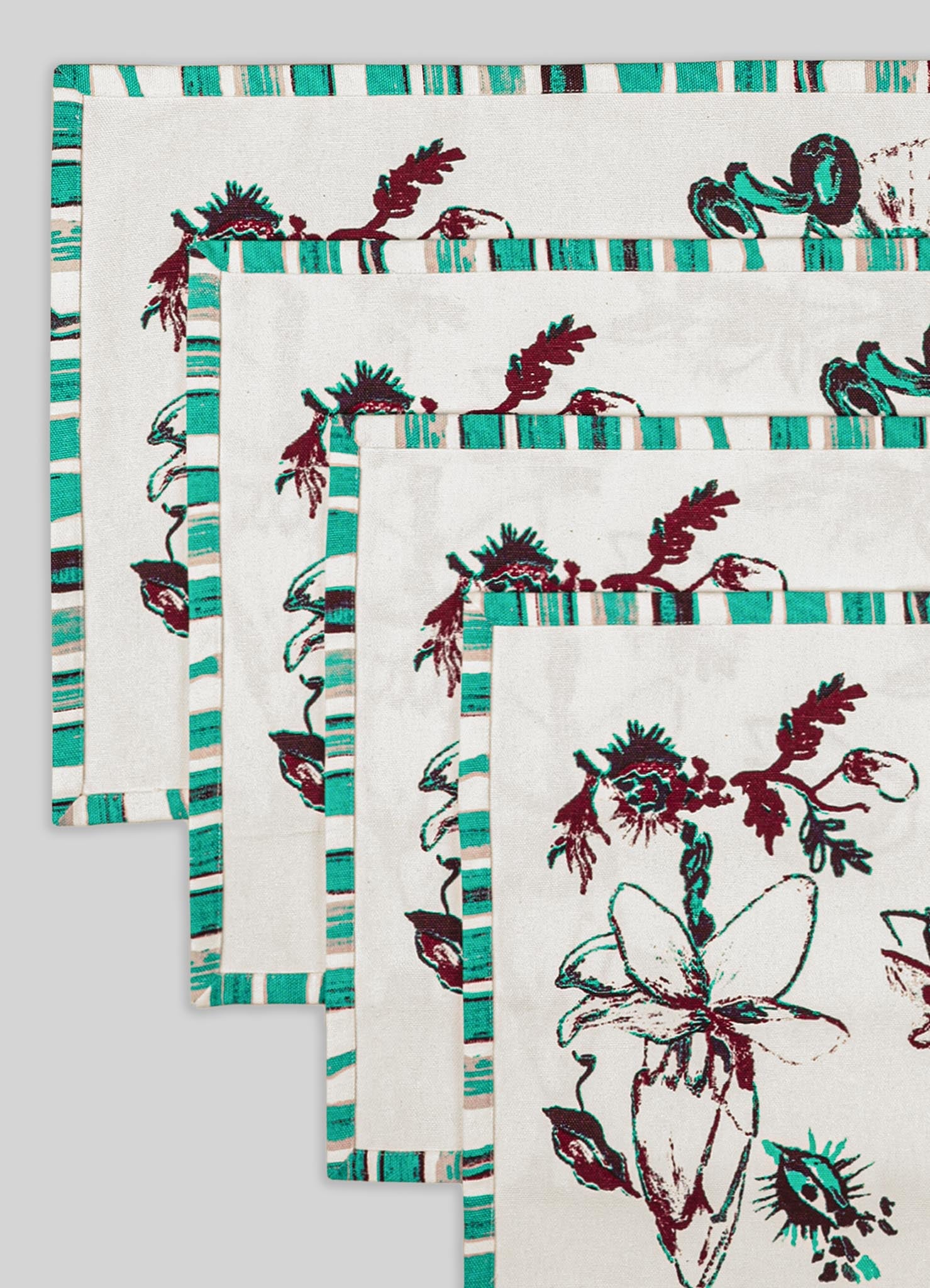 Fabric placemats with botanical floral motifs in dark red and teal, bordered by striped teal and beige edges, made of woven linen or cotton.