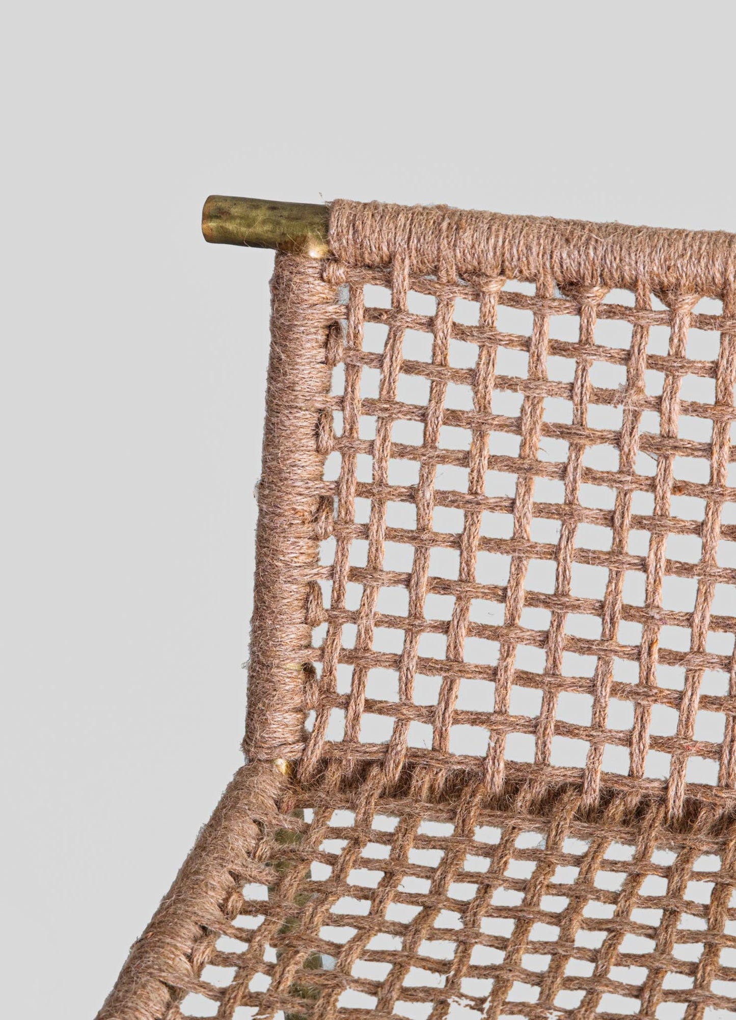 Close-up of a woven rattan chair with a natural beige finish, featuring an open grid pattern and a brass metal frame at the corner. The style appears rustic with handcrafted textile details.