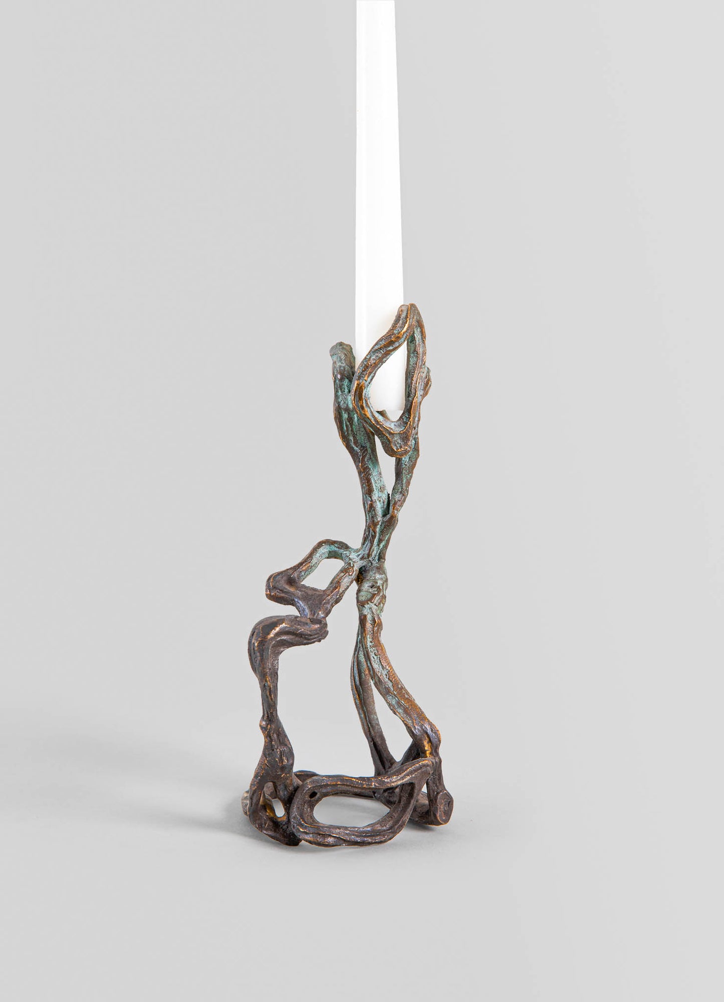 A sculptural candle holder made of metal with a rustic, textured finish in bronze tones, featuring organic, twisted shapes resembling driftwood. It holds a tall white taper candle and has a contemporary, handcrafted style.