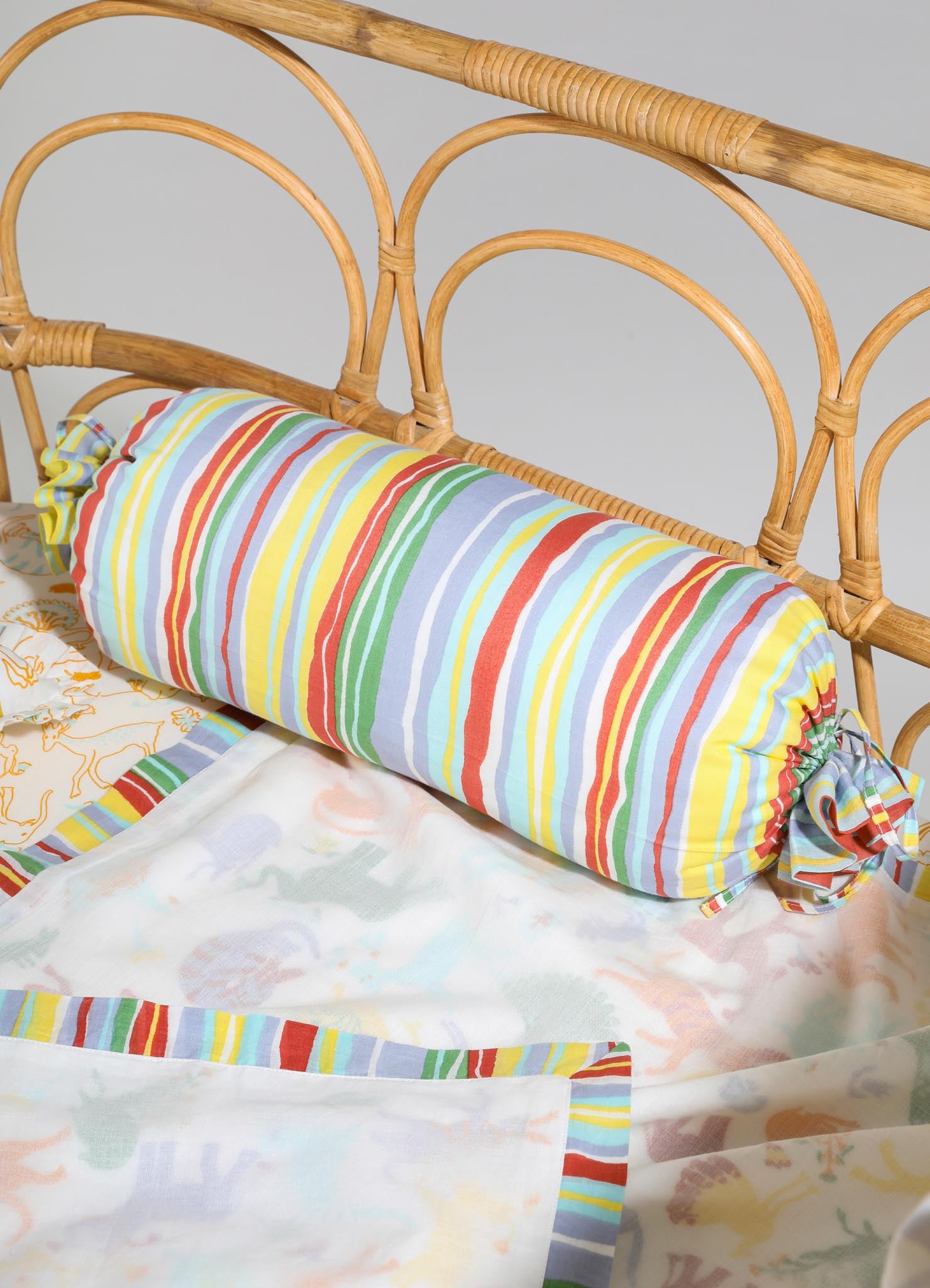Colorful striped fabric bolster pillow with ruched ends and tied bows on a rattan bed frame, paired with a light-colored bedspread featuring playful prints and matching striped accents.