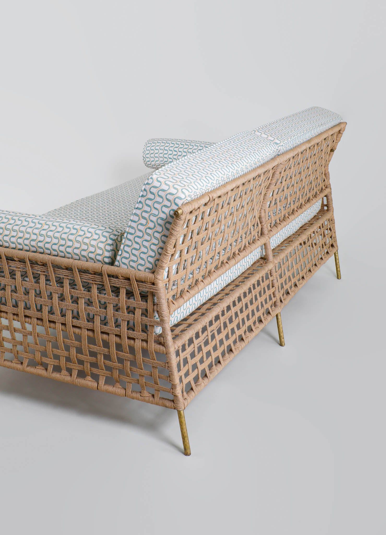 A woven rattan sofa with a lattice design, featuring brass legs and a padded cushion with a geometric pattern in neutral tones. The sofa has a matching backrest cushion and a decorative bolster pillow, presented in a minimalist setting.