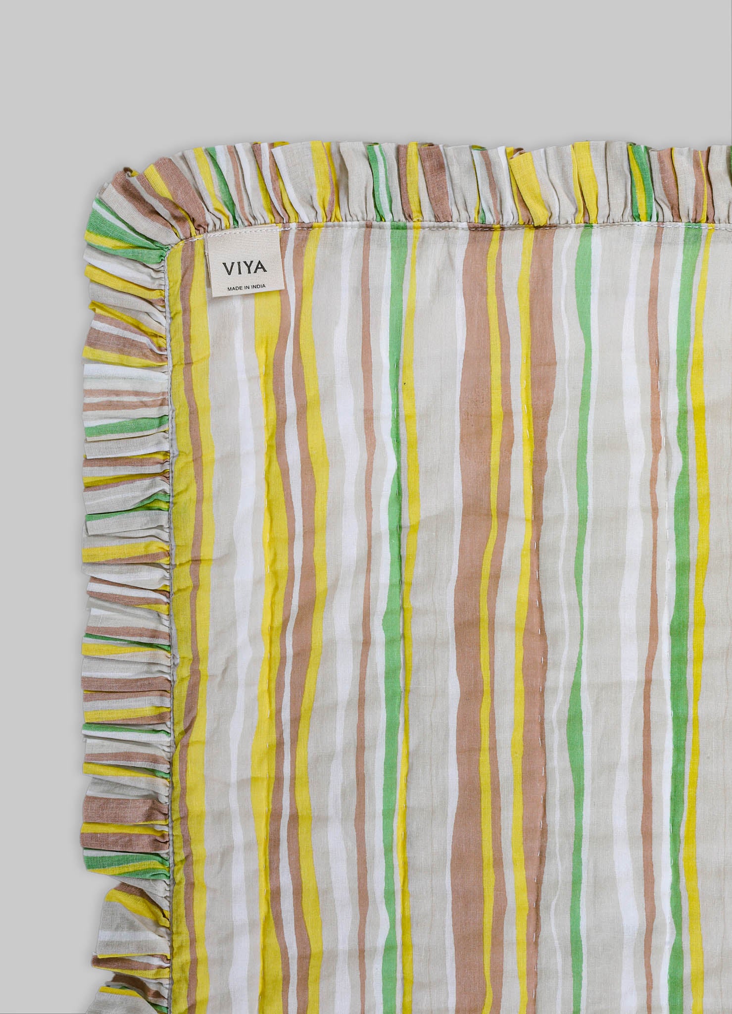 Striped cotton table runner with multicolored vertical lines in yellow, green, beige, and white, featuring a ruffled edge with a contrasting striped pattern. The label "VIYA" indicates it is made in India.