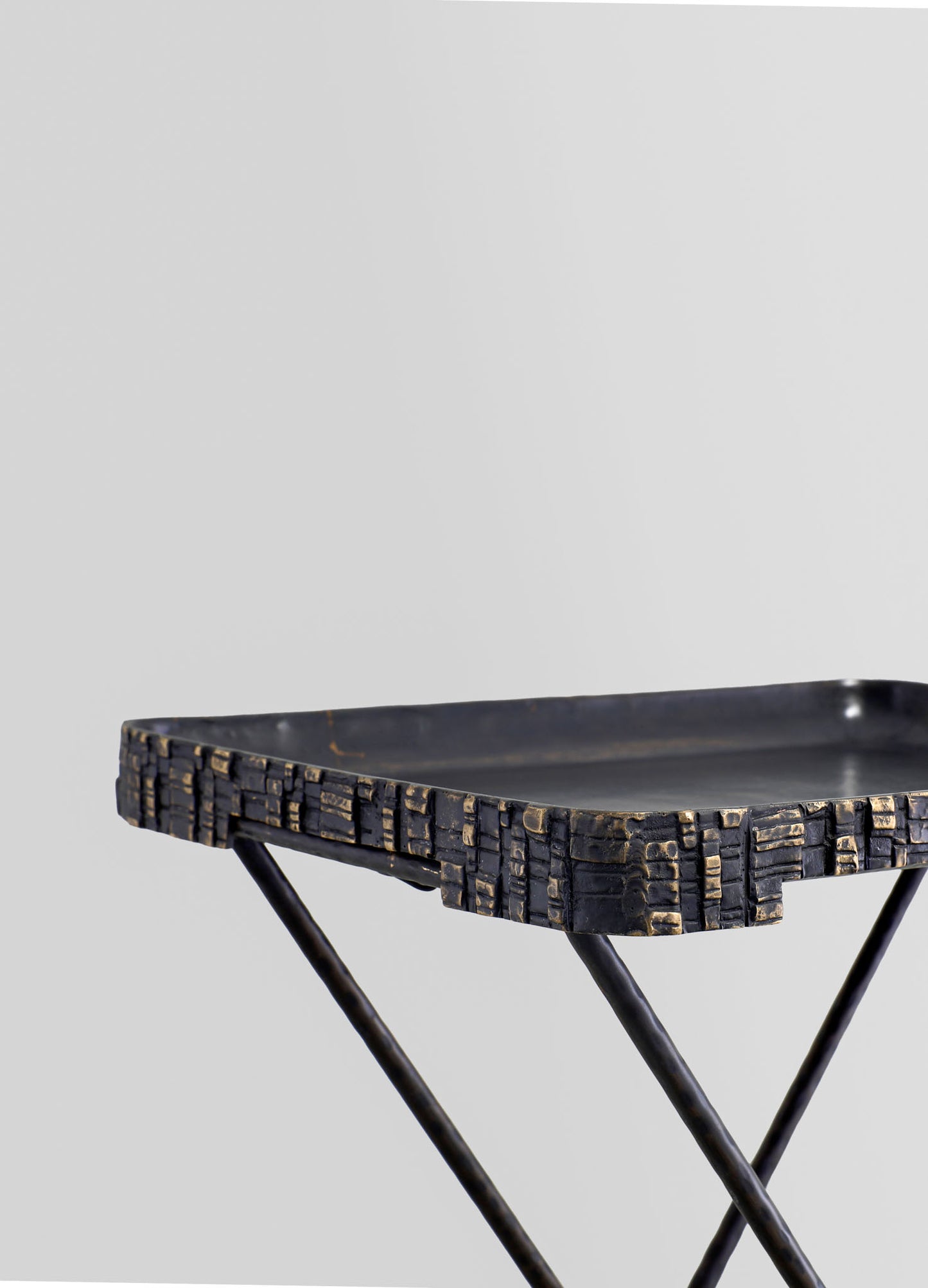 A black metal serving tray with textured, embossed geometric patterns on the edges, placed on a stand with slender, intersecting legs against a plain background.