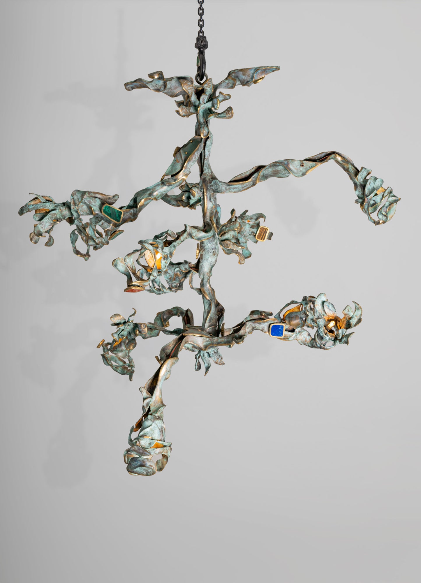 A sculptural chandelier made of metal with an organic, leaf-like design and a weathered, oxidized finish. The fixture features multiple small, colorful glass accents and illuminated bulbs, creating a contemporary artistic style.