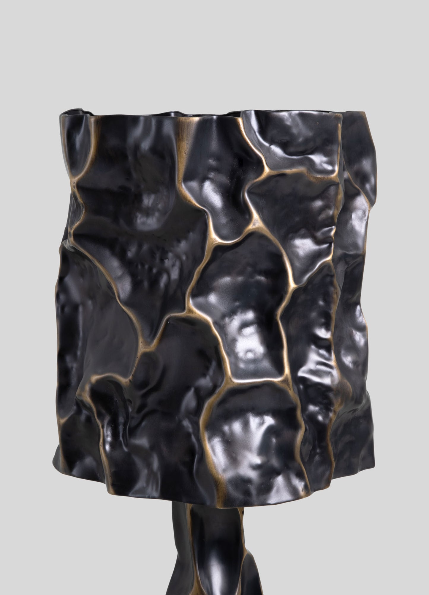 A decorative table lamp with a sculptural, irregular surface in black with gold accents, featuring a textured, organic motif resembling cracked stone or lava. The lamp has a contemporary style with a matte finish and an abstract, artisanal design.
