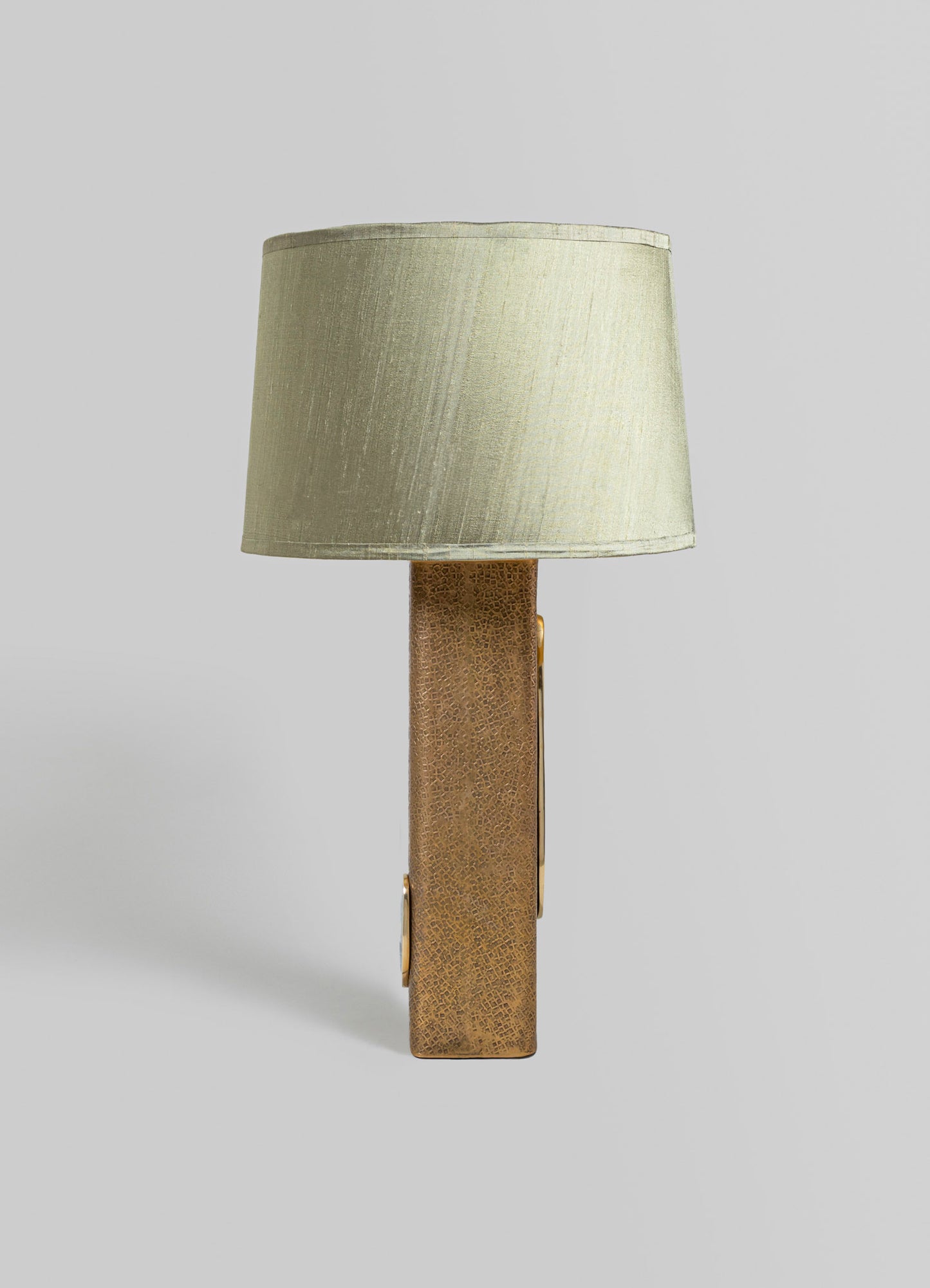 A table lamp featuring a cylindrical beige fabric lampshade and a rectangular base with a textured brass finish, designed in a contemporary style.