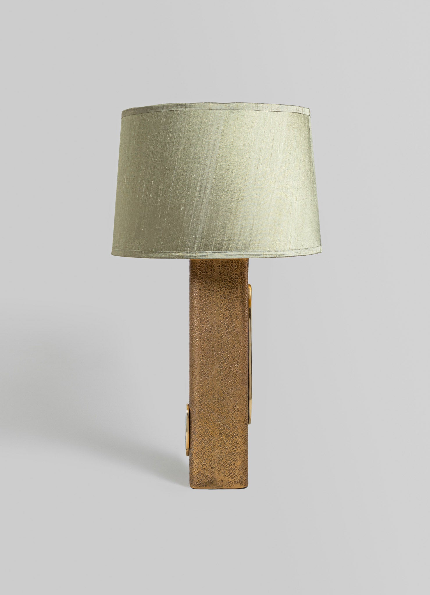 A table lamp featuring a cylindrical beige fabric lampshade and a rectangular base with a textured brass finish, designed in a contemporary style.