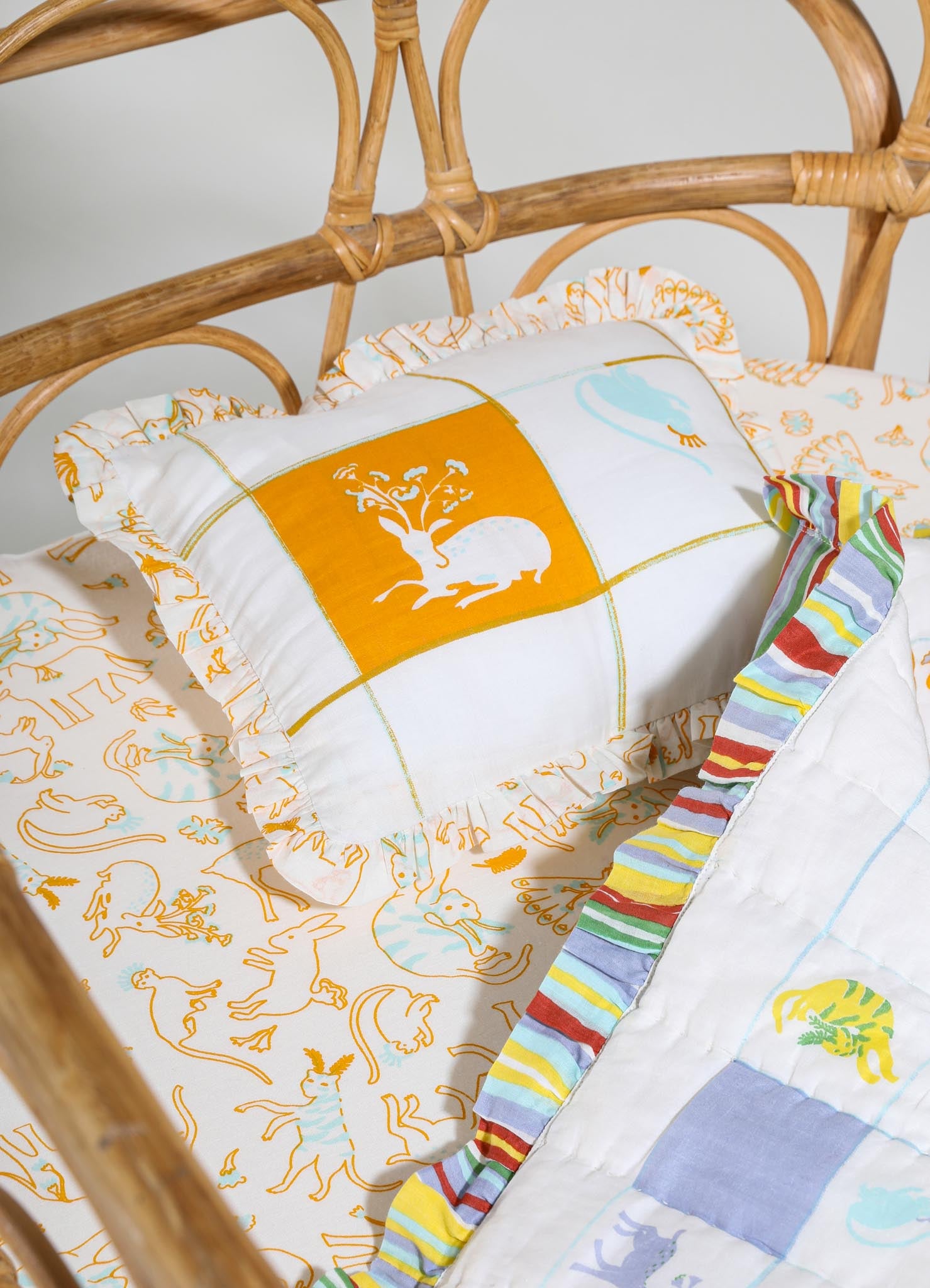 Cotton bed linen set with animal and botanical motifs in orange, yellow, and light blue, featuring ruffled edges and a multicolored striped border, displayed on a rattan bed frame. The design combines playful prints with a rustic, handcrafted style.