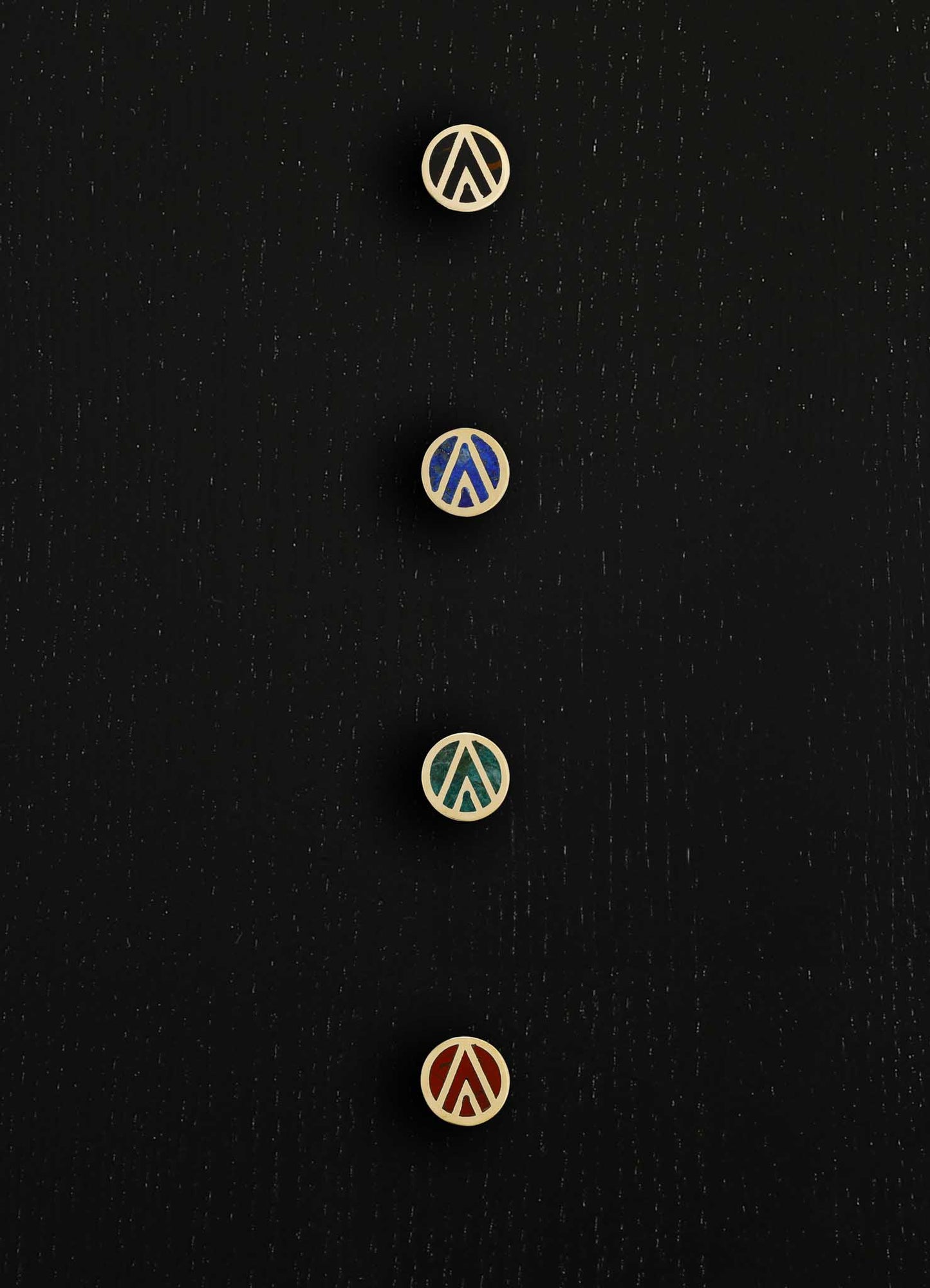 Four round metal buttons with cut-out geometric motifs, featuring a mix of matte metallic finishes in gold, blue, green, and red, arranged vertically on a black textured background.