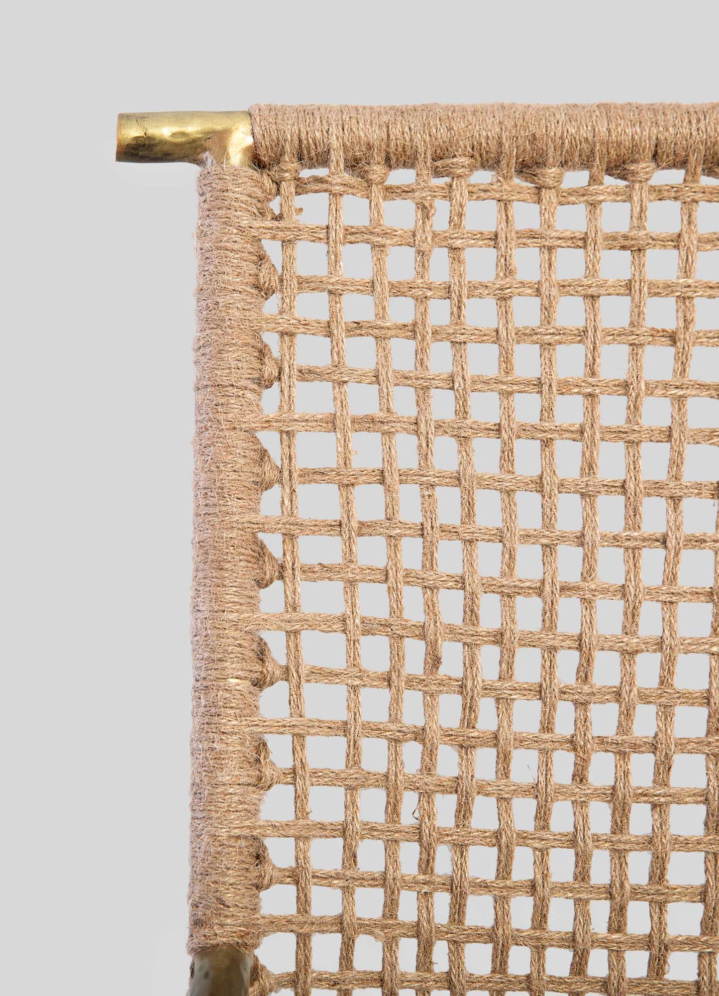 Close-up of a woven rattan or cane chair back with a grid pattern, featuring natural beige tones and a thin wooden frame. The top corner shows a brass or gold-colored metal piece attached to the frame for support.