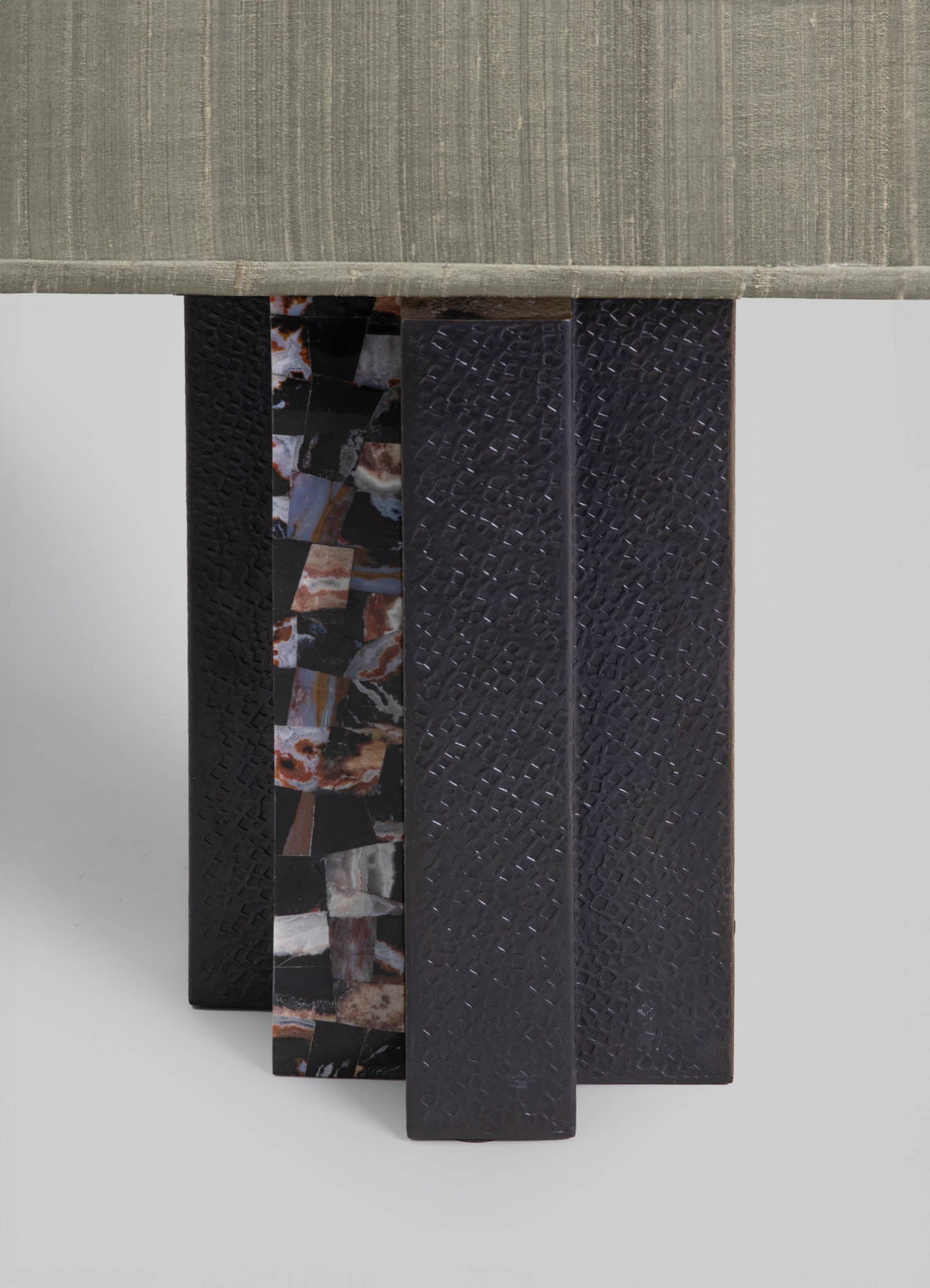 A table with a beige fabric top and a base made of decorative panels, including one with a multicolored marble pattern and others with textured black surfaces. The design combines contemporary style with mixed materials and patterns.