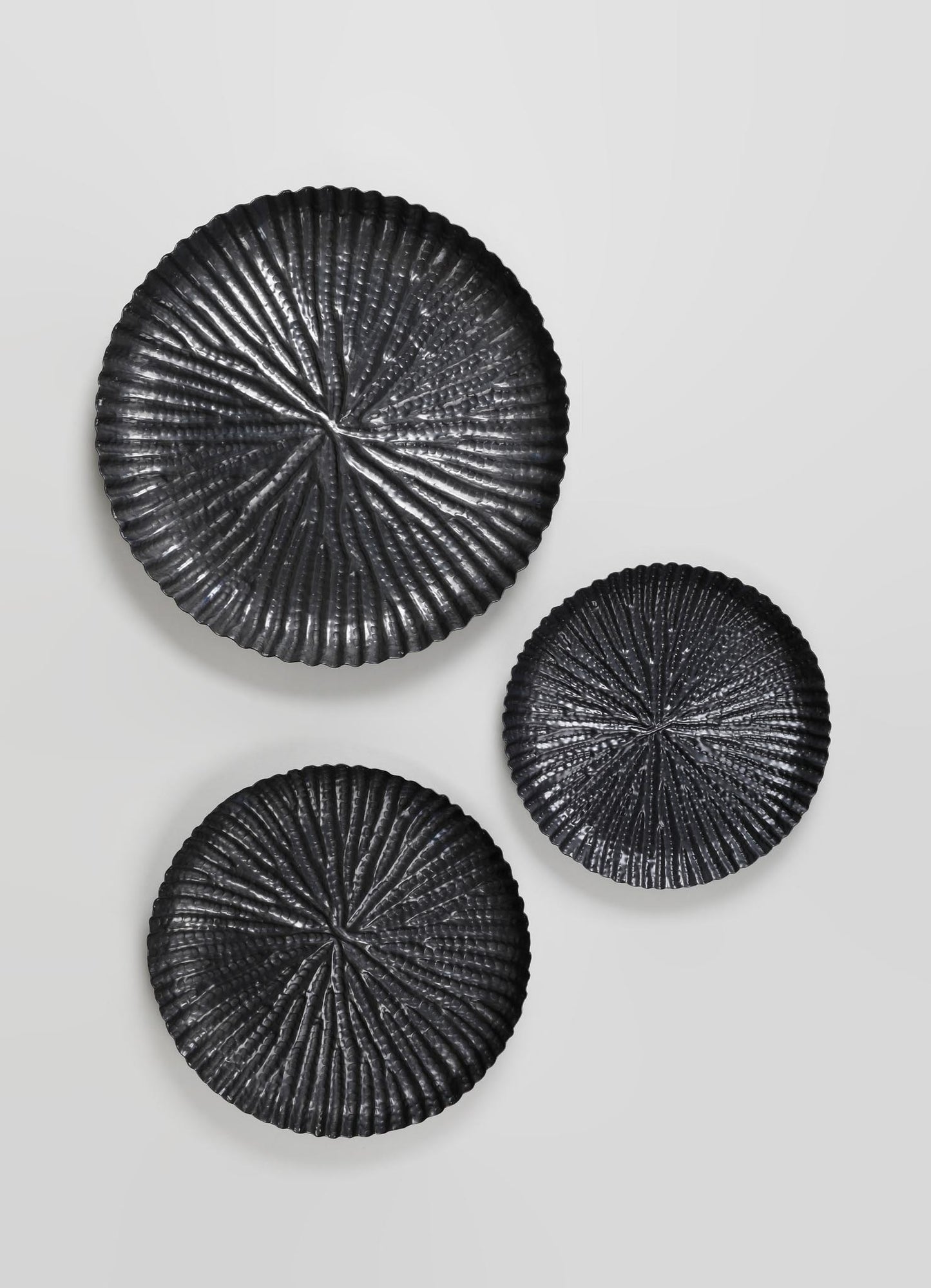 Set of three round wall decor pieces made of black metal with textured, radiating patterns. The designs resemble abstract, leaf-like or floral motifs with a glossy finish, arranged on a plain light-colored wall.
