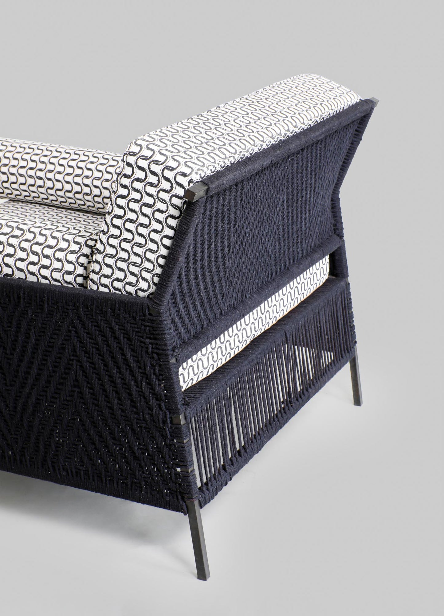 A woven rattan armchair with a black finish, featuring a geometric pattern on the back and sides, and a cushion with black and white abstract wave motifs. The chair has a contemporary style with slender metal legs.