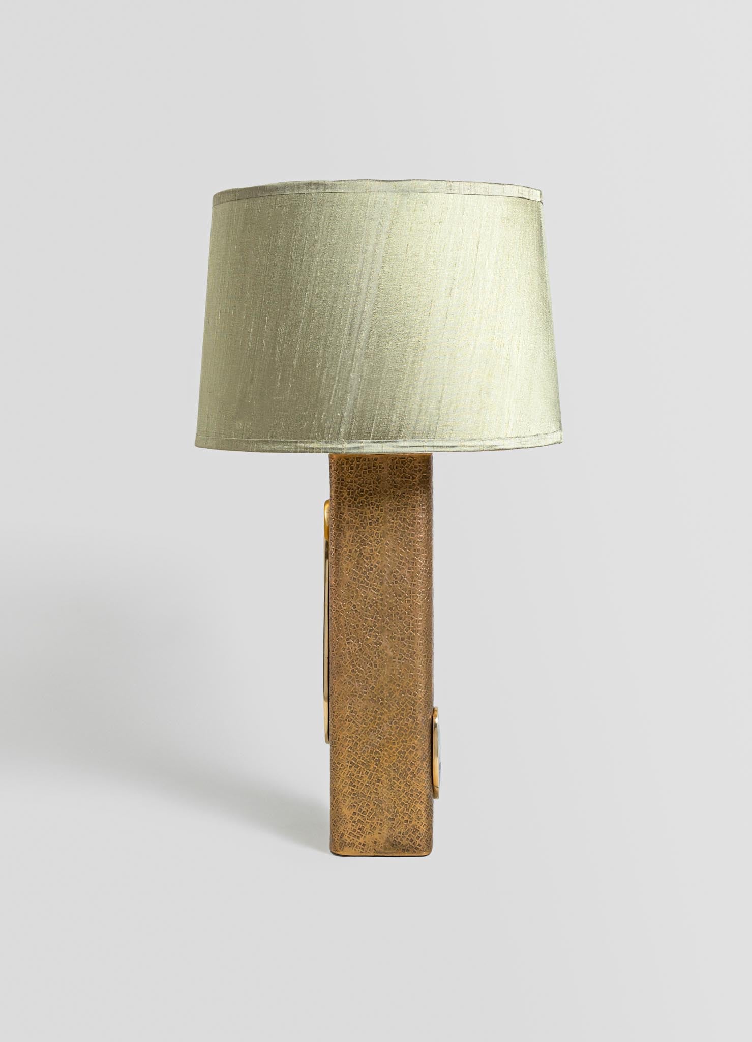 A table lamp with a textured brass base featuring a rectangular shape and metallic finish, topped with a cylindrical beige fabric lampshade.