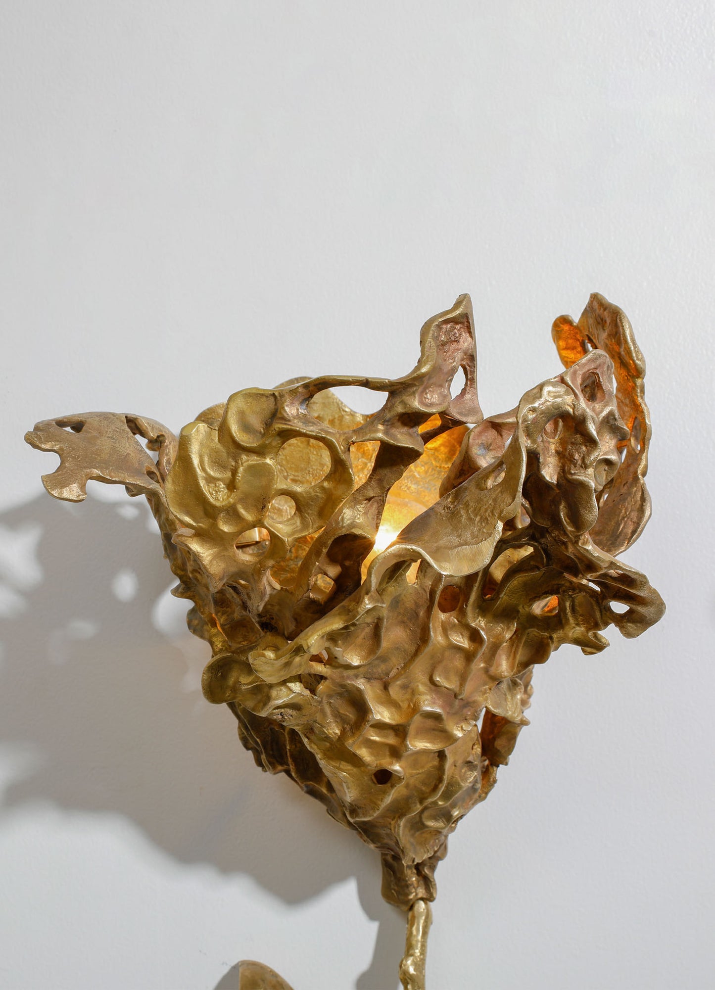 A sculptural table lamp with an organic, abstract form made of textured metal in a gold finish, with intricate hollowed-out patterns resembling natural coral or mineral formations. The lamp is placed against a plain white wall, casting dynamic shadows.