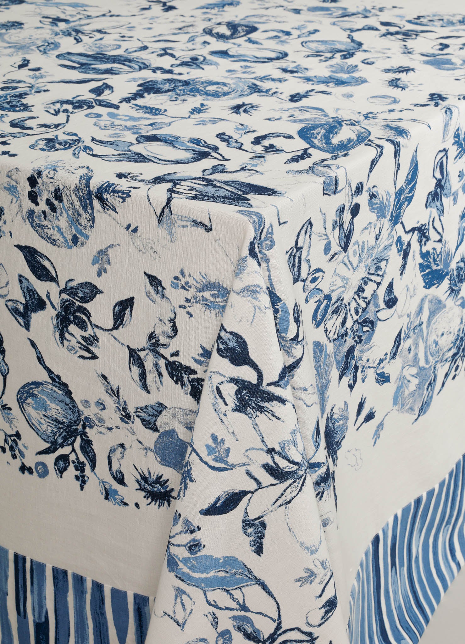 Tablecloth with a floral and botanical pattern in shades of blue on a white background, crafted from fabric material. The edges feature a blue and white striped border, creating a casual yet elegant style.