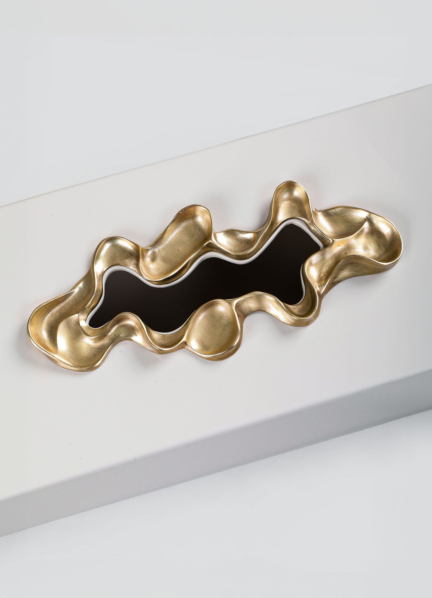 A decorative mirror with an abstract, wavy-shaped frame in metallic gold finish, mounted on a white wall. The mirror has irregular, organic contours that evoke a modern, sculptural style.