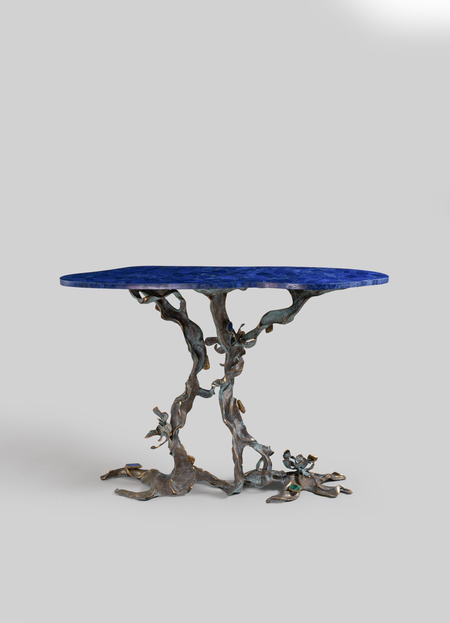 A round table with a blue mosaic tabletop and an irregular, sculptural metal base with a weathered finish, combining organic and contemporary design elements.