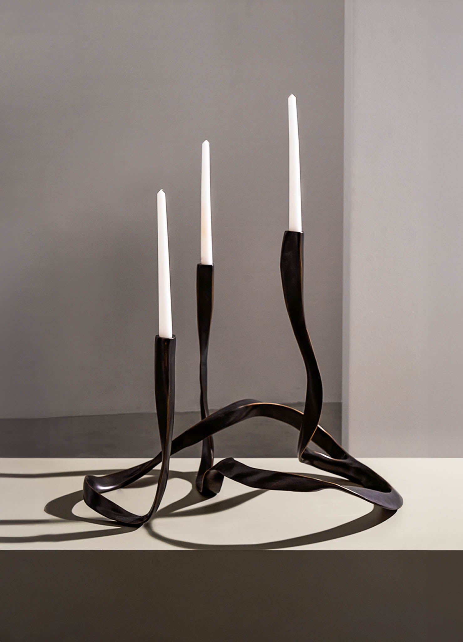 A sculptural candle holder with a dark, twisted metal design supporting three tall, white taper candles, set against a neutral background. The piece has an organic, contemporary style with flowing, irregular shapes.
