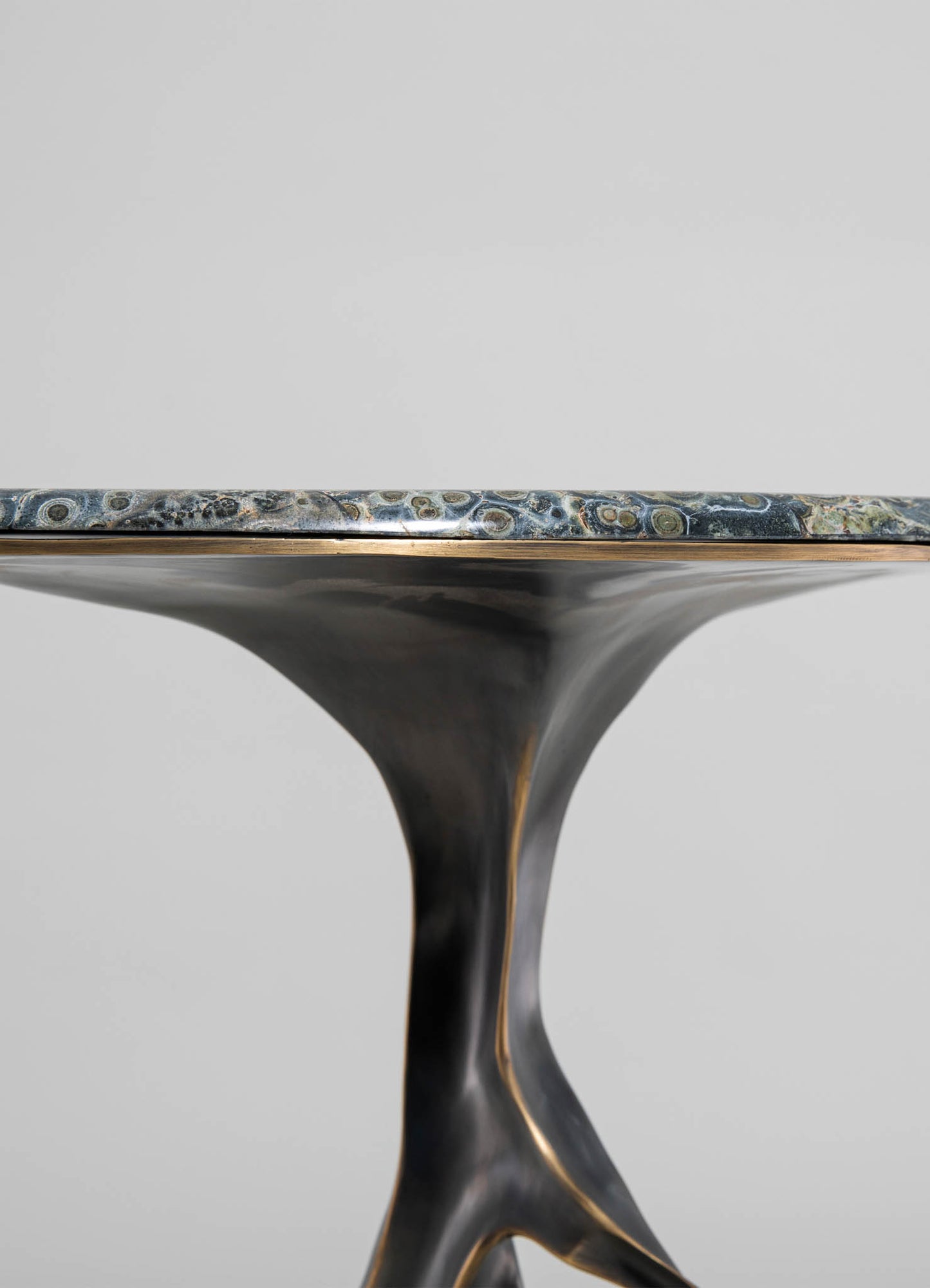 A close-up of a modern side table featuring a sculptural, curved metal base with a matte black finish and a polished, multi-toned marble top with swirls of gray, green, and beige.
