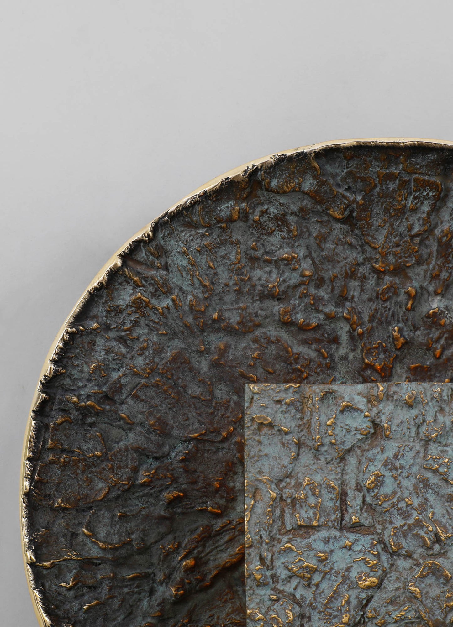 A round decorative tray with a textured, oxidized metal surface in dark brown and blue-gray tones, edged with a thin brass rim. The tray has a rustic, artisanal style with an uneven, distressed finish.