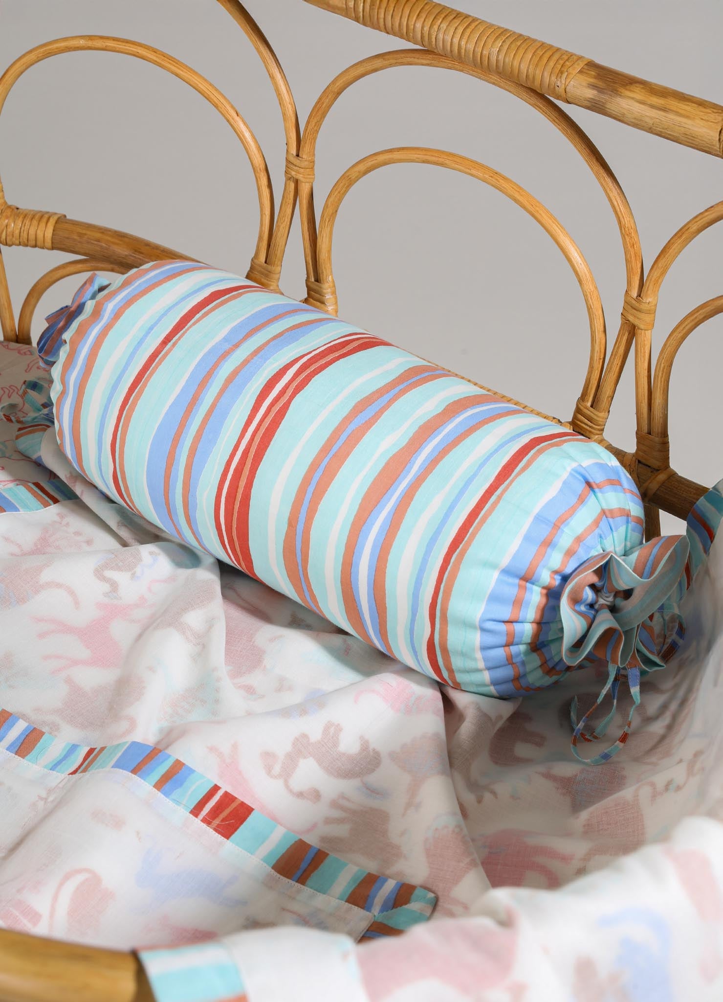 Striped bolster pillow with multicolored lines in shades of red, blue, and white, resting on a bed with a bamboo headboard. The bed features light pink and beige patterned bedding with floral and animal motifs.