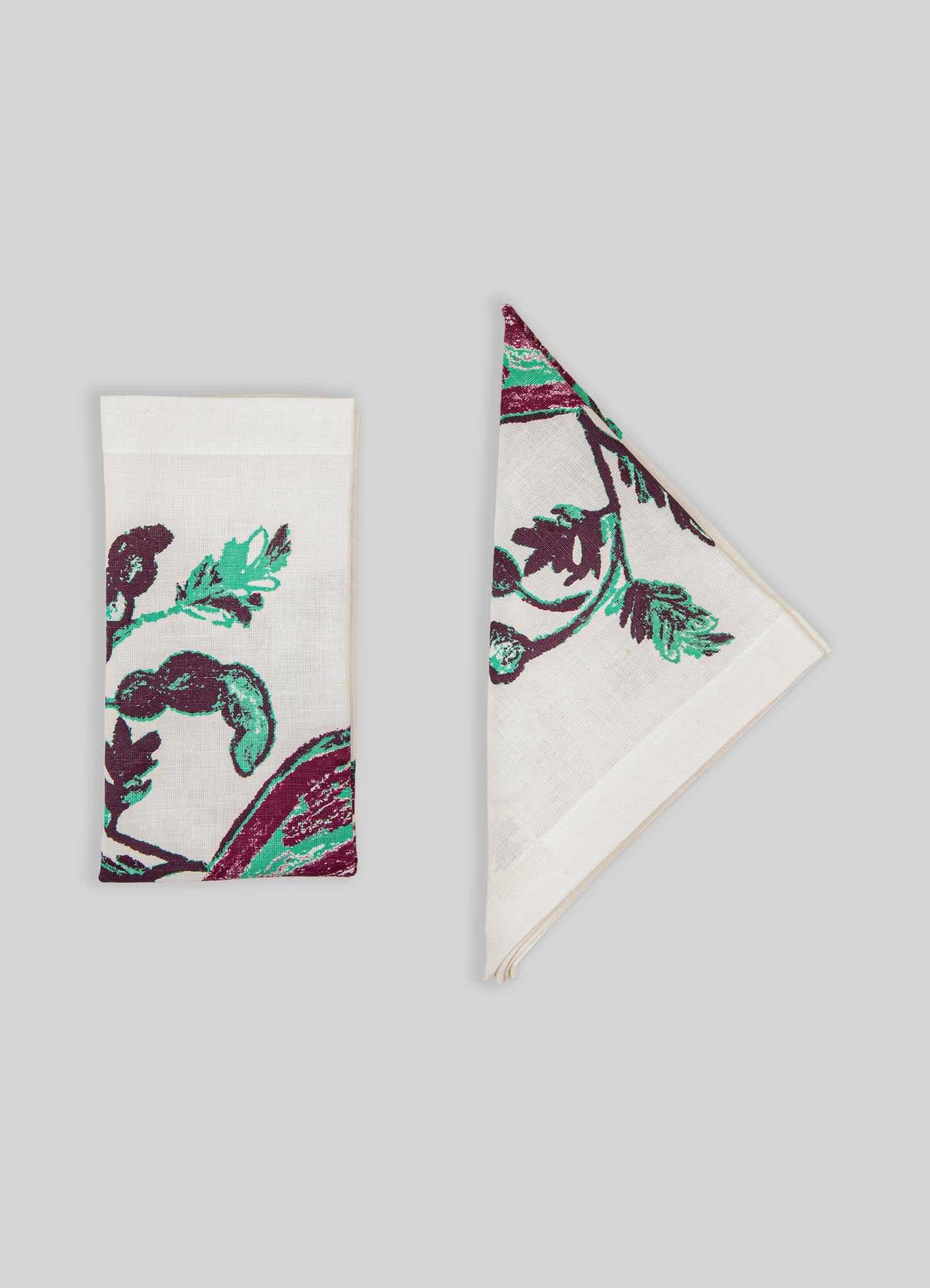 Cotton napkins with a floral motif in shades of green and purple, featuring a textured fabric finish. One napkin is folded flat, while the other is partially unfolded, displayed against a plain light gray background.