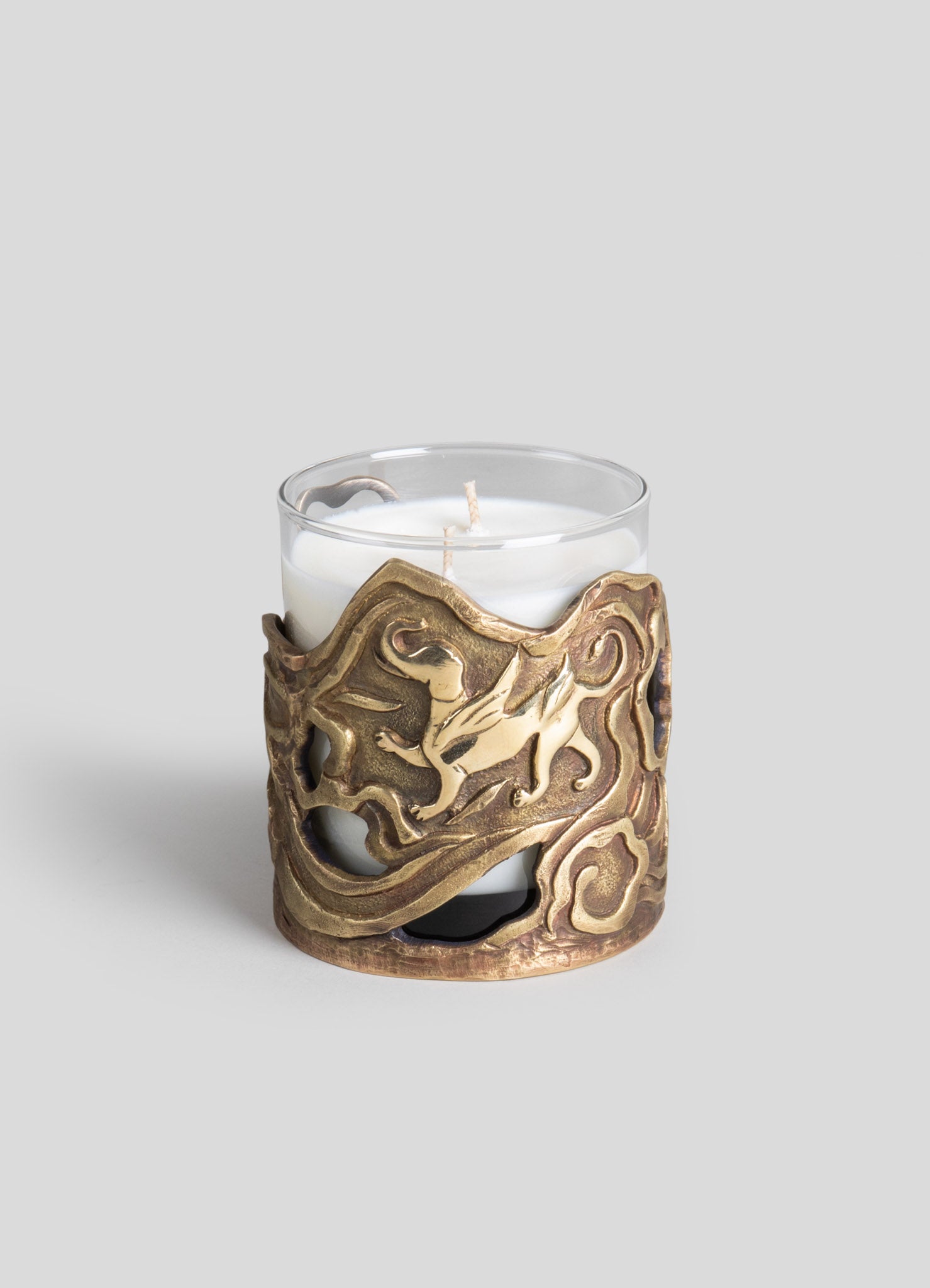 Glass candle holder with an ornate brass or bronze holder featuring a carved dragon motif, designed to encase a white pillar candle. The decorative metalwork has a textured, antique finish.