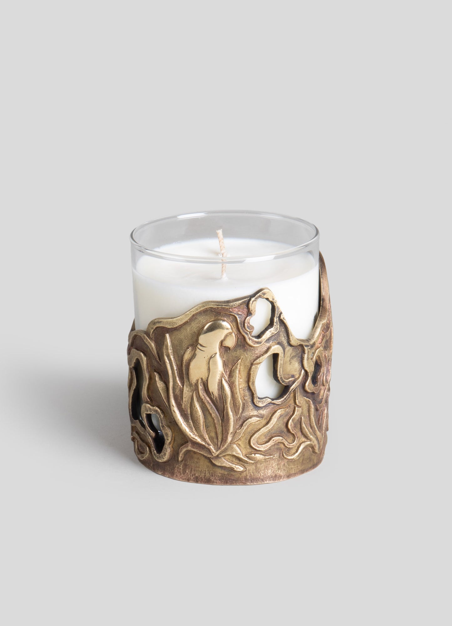 A white pillar candle inside a glass holder with a brass or metallized bronze decorative cover featuring organic, flowing motifs.