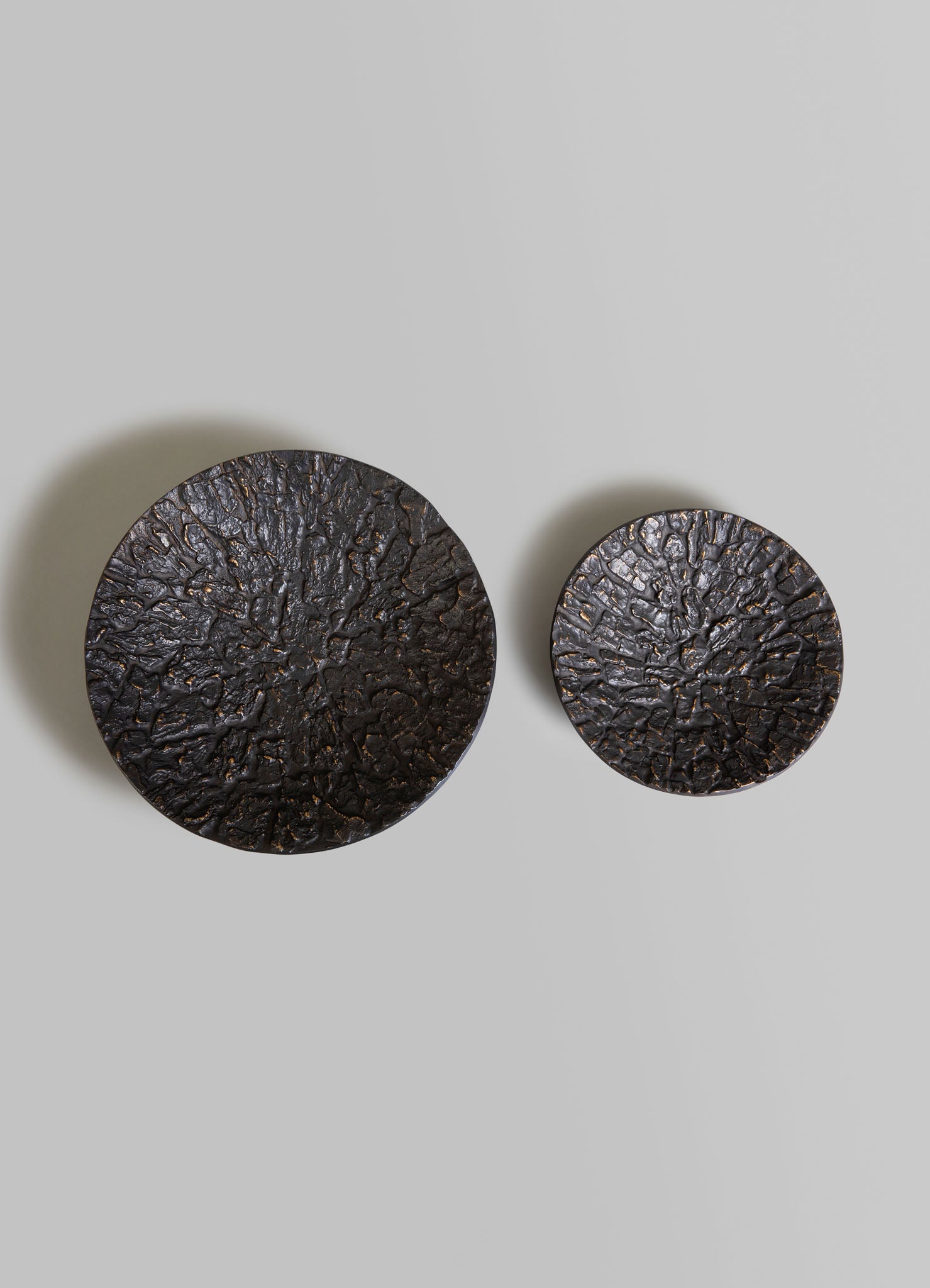 Two round decorative wall hangings with textured surfaces resembling cracked or fissured patterns in dark metallic tones, mounted on a light gray wall. The pieces have an organic, rustic style with a rough, uneven finish.